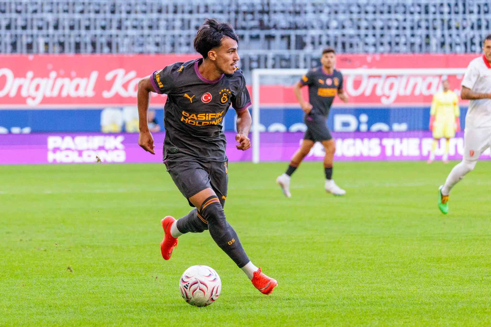 Junger Galatasaray-Spieler dribbelt mit roten Stollenschuhen über den Rasen, Ball am linken Fuß.