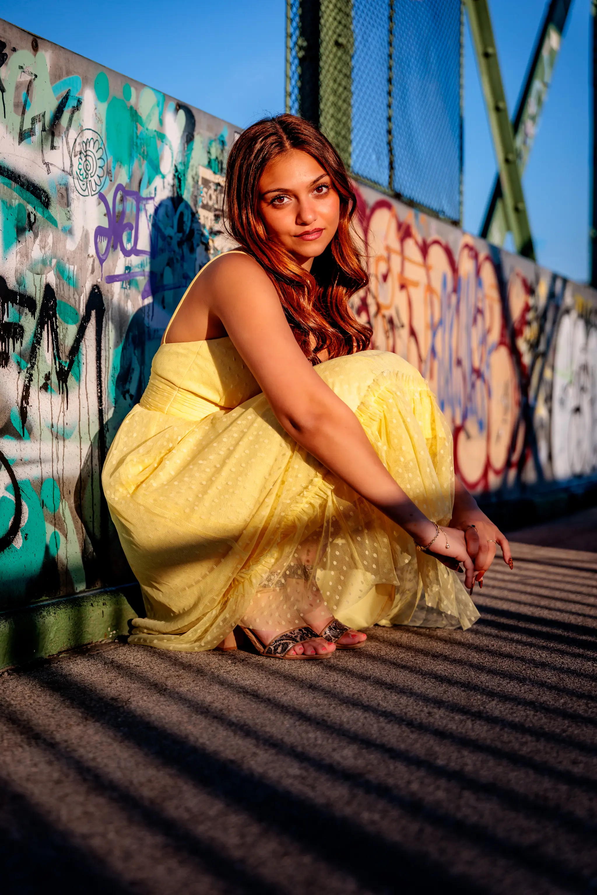 Junge Frau in gelbem Kleid kauert vor Graffiti-Wand mit Sonnenlicht und Schatten. 