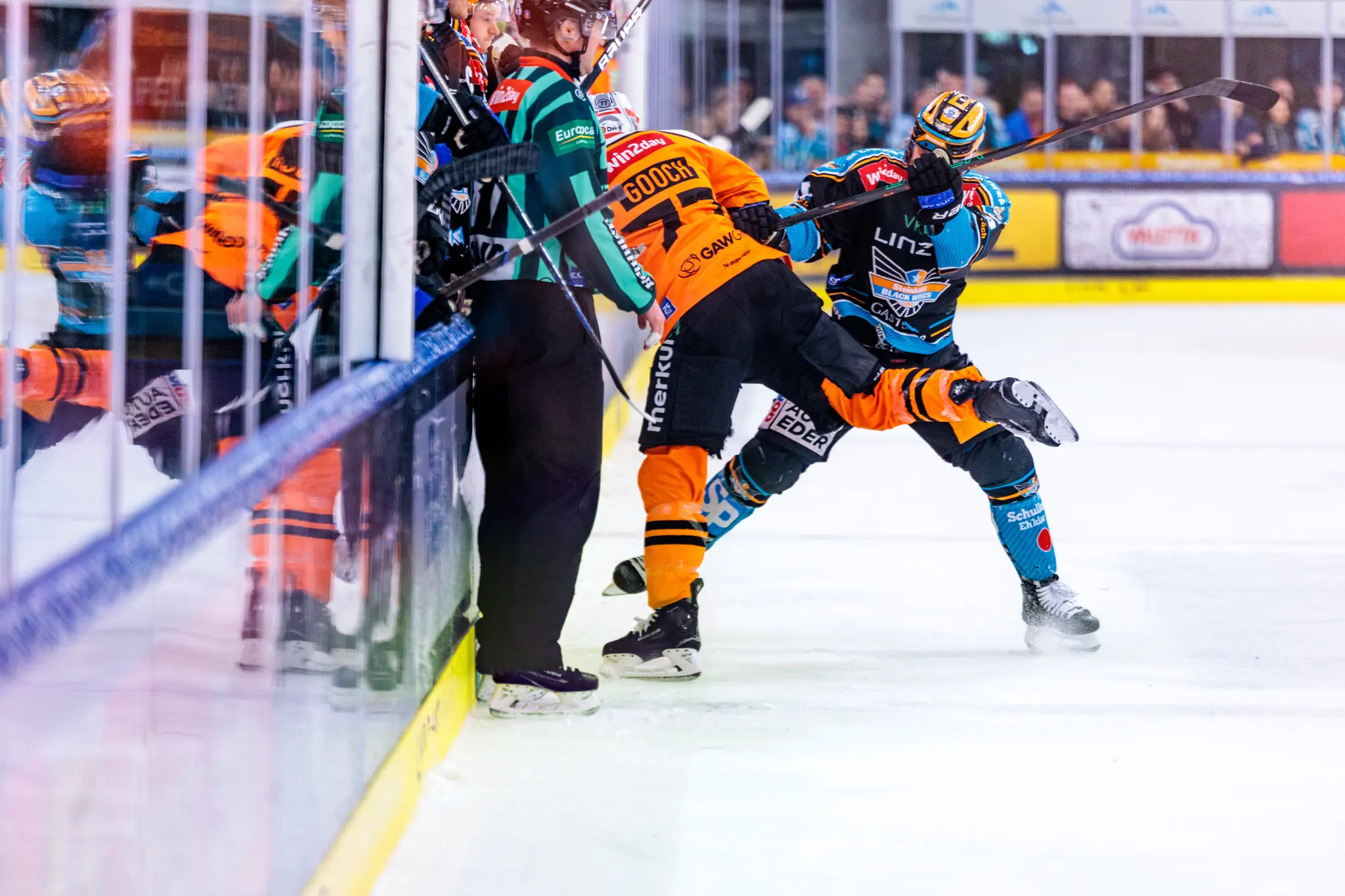 Ein Eishockeyspieler der Black Wings Linz in schwarz-türkisem Trikot und ein orange gekleideter Gegenspieler der Grazer 99ers sind intensiv im Kampf um den Puck an der Bande 