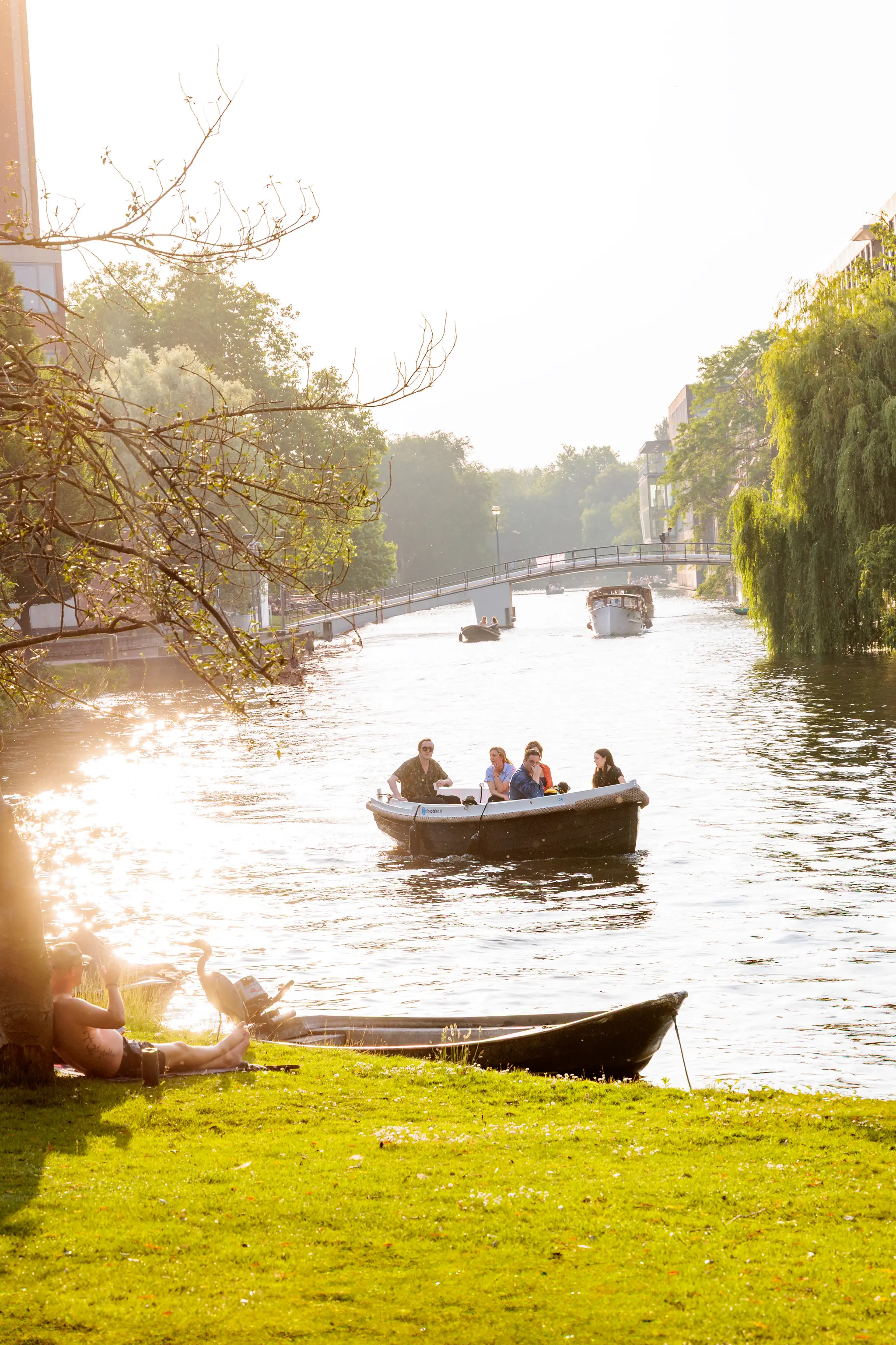 Ein Boot mit Personen gleitet auf einem sonnenbeschienenen Kanal, während am Ufer ein Mann entspannt und ein Reiher danebensteht 