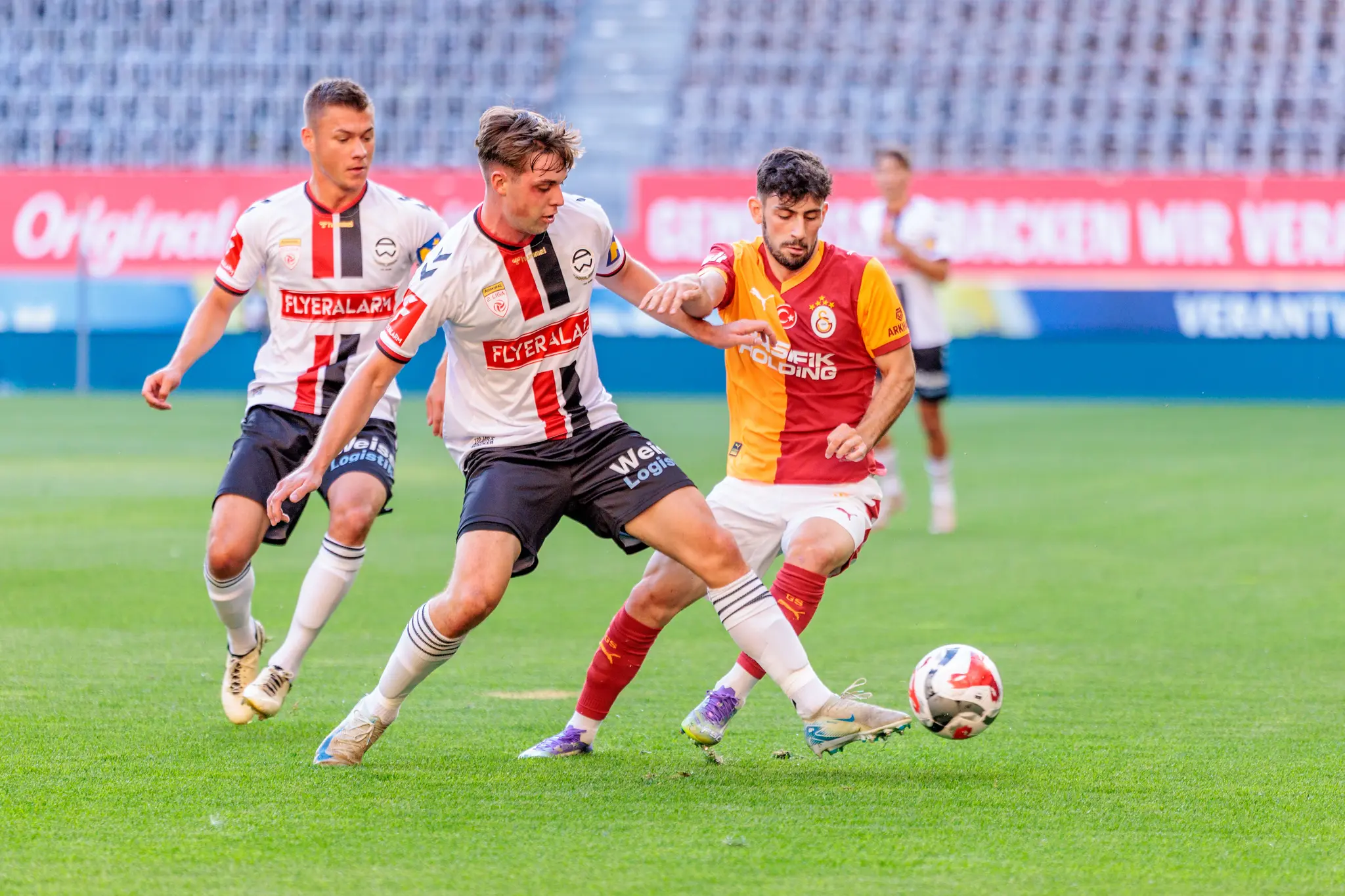 Zwei Fußballspieler in Trikots mit weiß-rot-schwarzen beziehungsweise rot-orangen Streifen liefern sich einen Zweikampf um den Ball 