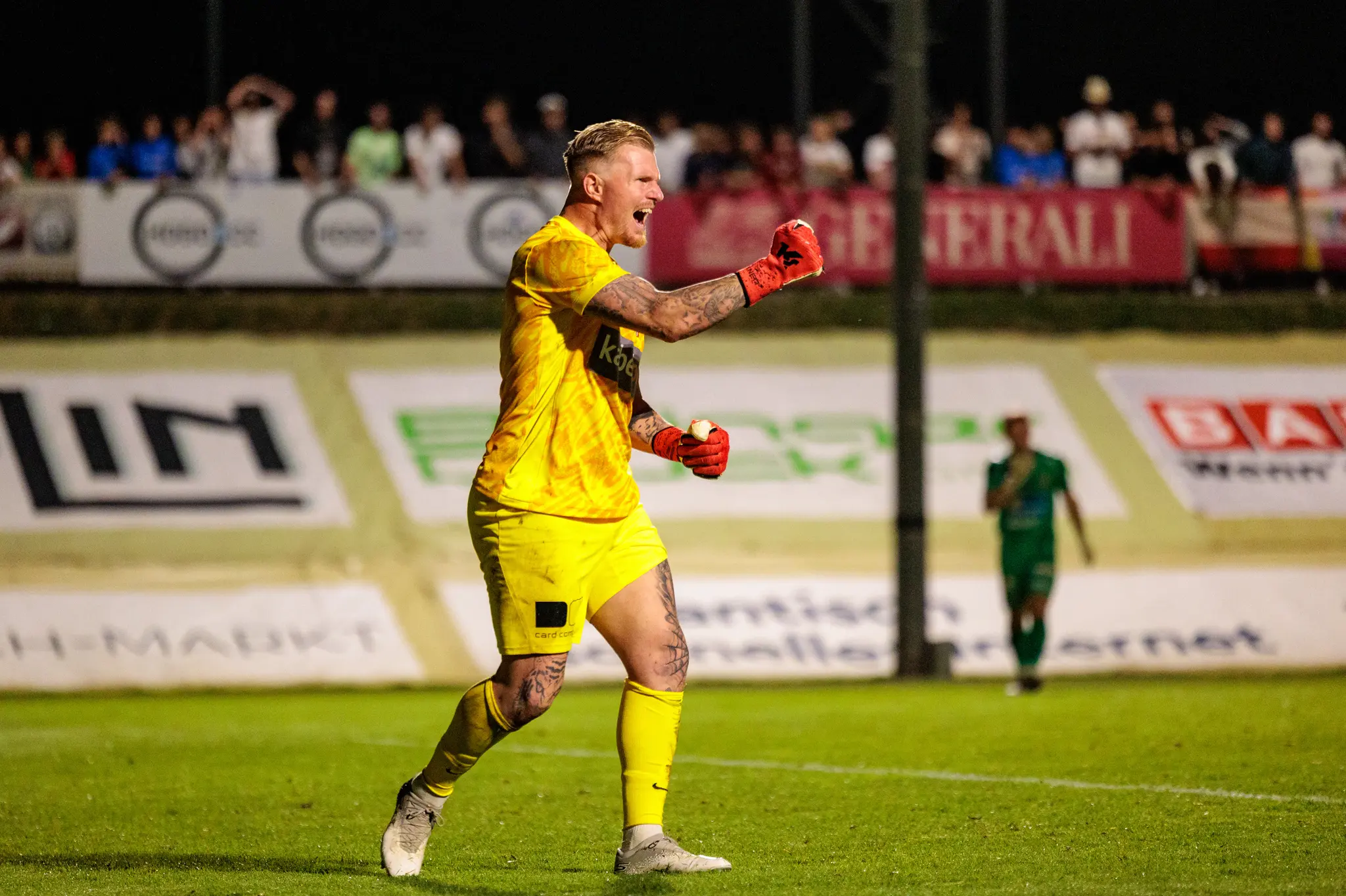 Ein leidenschaftlich jubelnder Fußballtorwart in leuchtend gelbem Trikot und roten Handschuhen feiert mit geöffnetem Mund und geballten Fäusten auf dem Spielfeld nach einem Elfmeterschießen