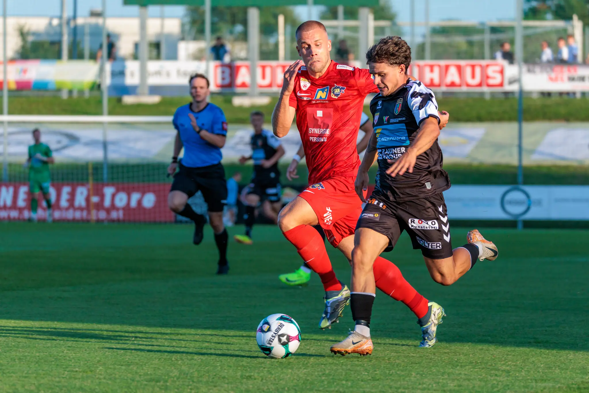 Ein Fußballspieler von SKN St. Pölten im roten Trikot und ein Gegner der SPG HOGO Wels im schwarzen Trikot bestreiten einen hitzigen Zweikampf um den Ball