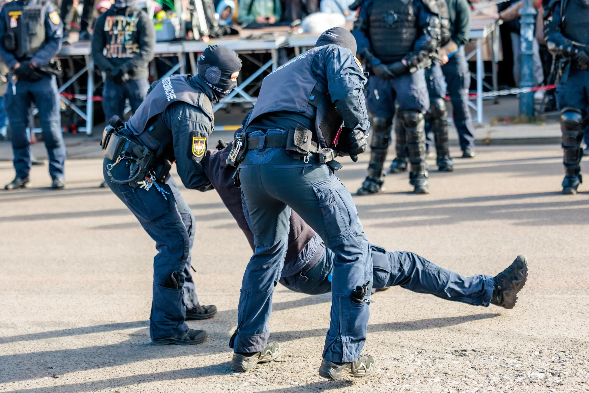Zwei Beamte der Justizwache in dunkler Einsatzausrüstung beugen sich über eine am Boden liegende Person, weitere Beamte mit Schutzhelmen und Schilden stehen im Hintergrund.