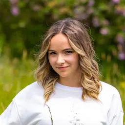 Eine junge Frau mit blonden Locken und einem weißen Pullover lächelt leicht in die Kamera vor einem natürlichen, unscharfen grünen Hintergrund lens: RF70-200mm F2.8 L IS USM