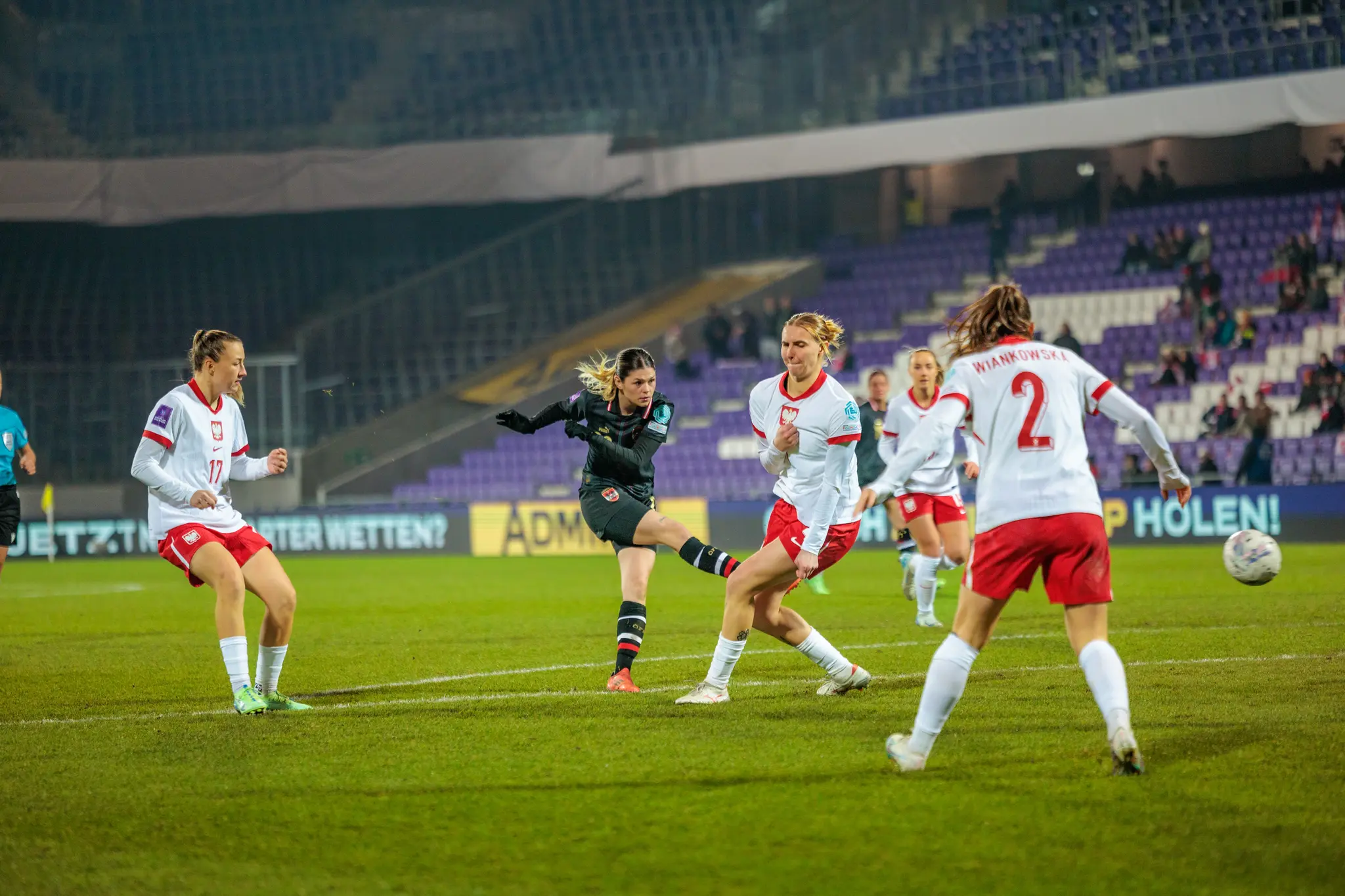 Eine österreichische Fußballerin im schwarzen Trikot tritt während eines internationalen Frauenfußballspiels den Ball gegen polnische Spielerinnen im weiß-roten Dress 
