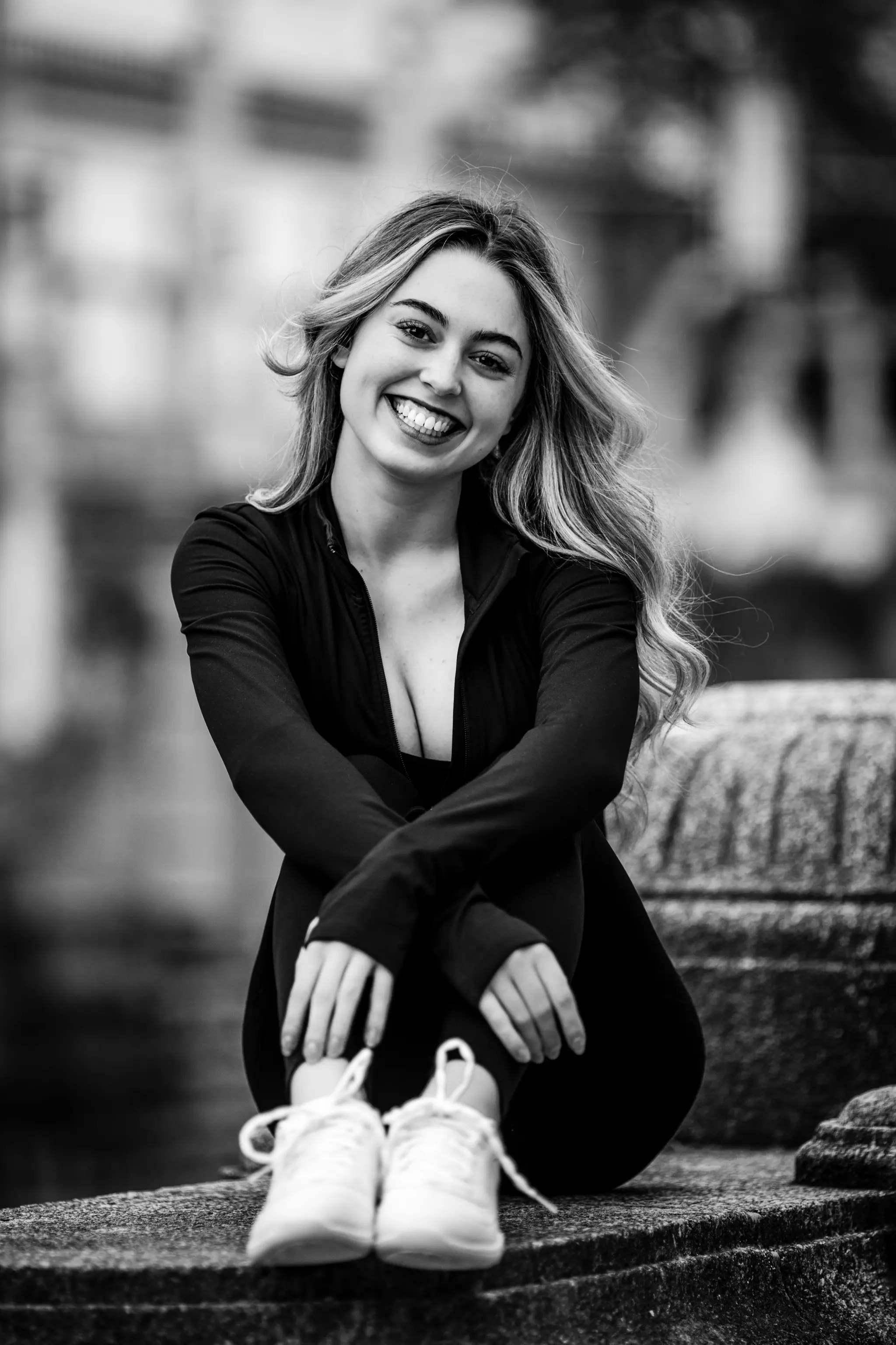 Ausdrucksstarkes Schwarz-Weiß-Porträt von Model Valentina, die in einem dunklen, sportlichen Outfit auf einer Steinmauer im Stadtpark Wien sitzt und herzlich lacht