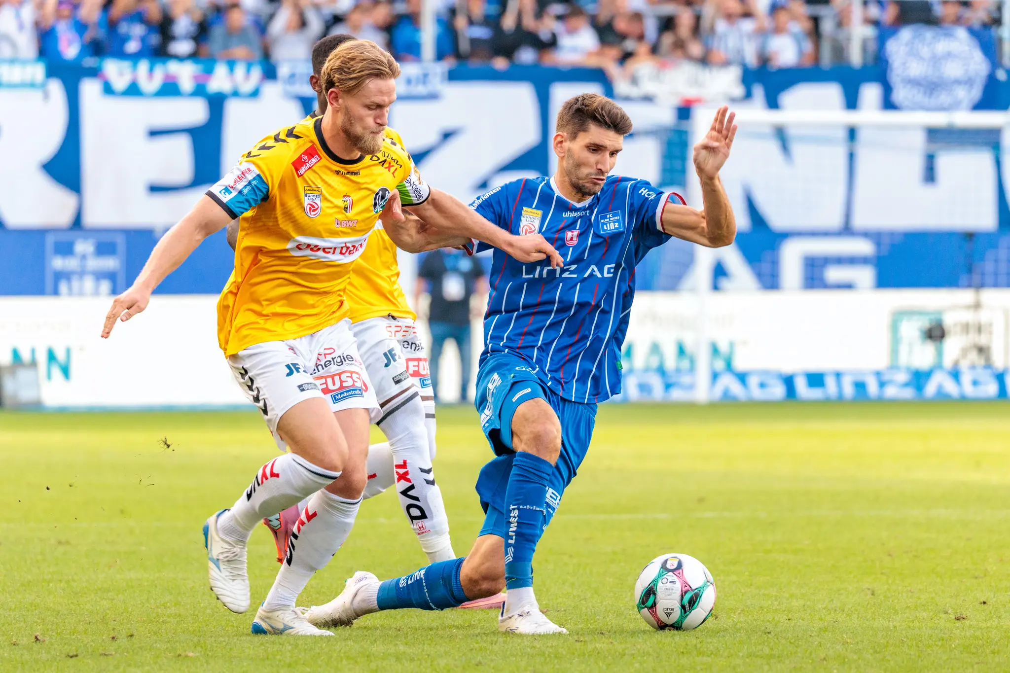 Ein Spieler des SV Ried im gelben Trikot setzt einen Spieler des FC Blau-Weiß Linz im blau-weiß gestreiften Trikot im Zweikampf um den Ball unter Druck