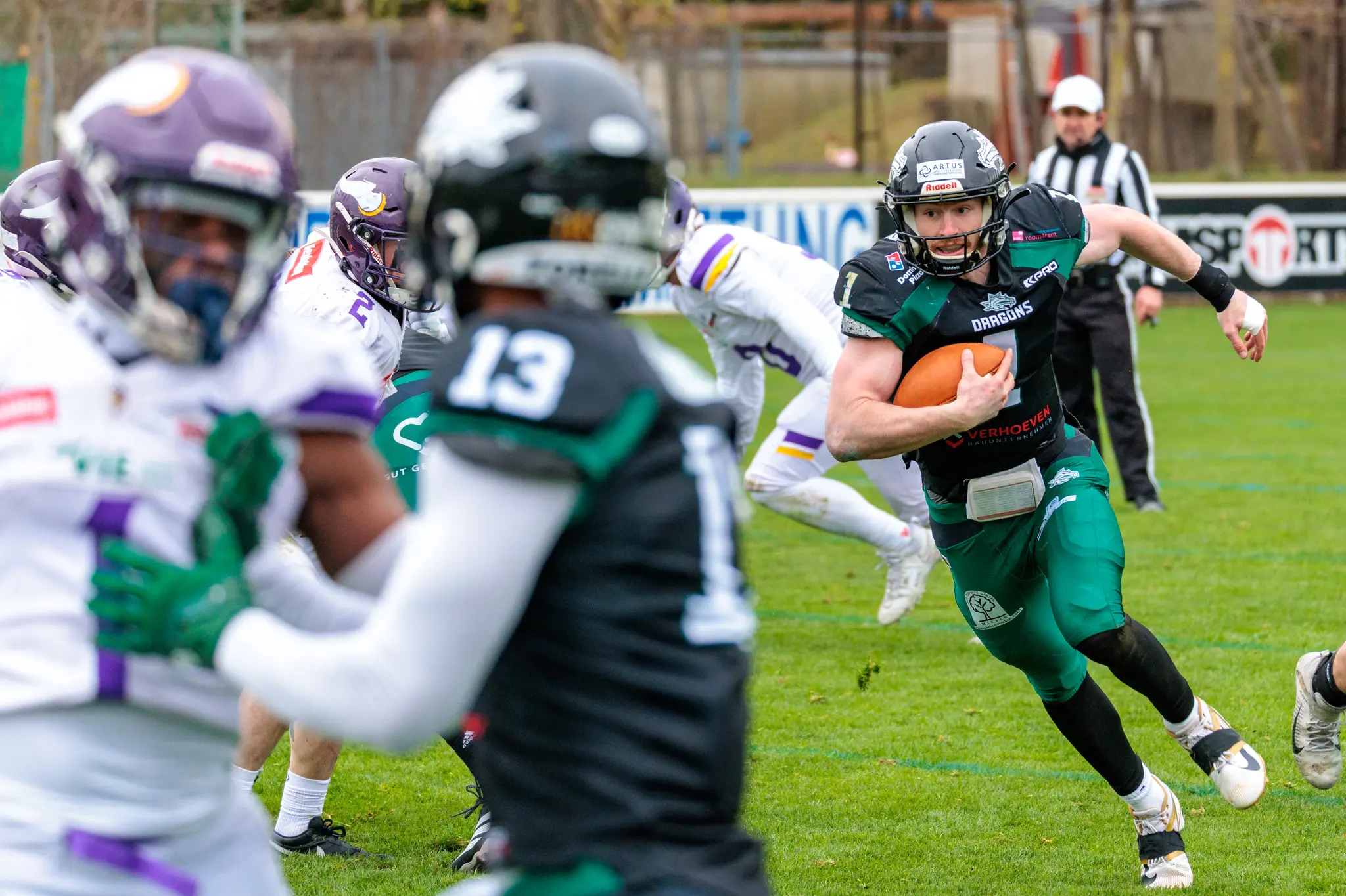 Ein American-Football-Spieler der Danube Dragons in grüner Uniform sprintet mit dem Football über das Feld, während unscharfe Spieler der Vienna Vikings im Hintergrund folgen