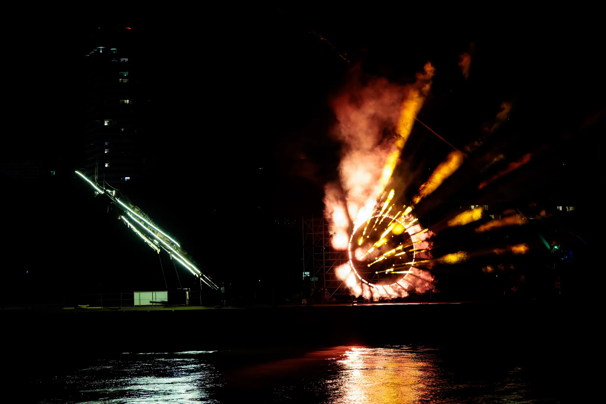 Nächtliche Lichtershow über Wasser während der Linzer Klangwolke 2024: Ein goldener Ring sprüht Funken und Rauch, umgeben von orangen Lichtstrahlen. Links eine lange, grüne Lichtrampe mit Spiegelungen im Wasser 