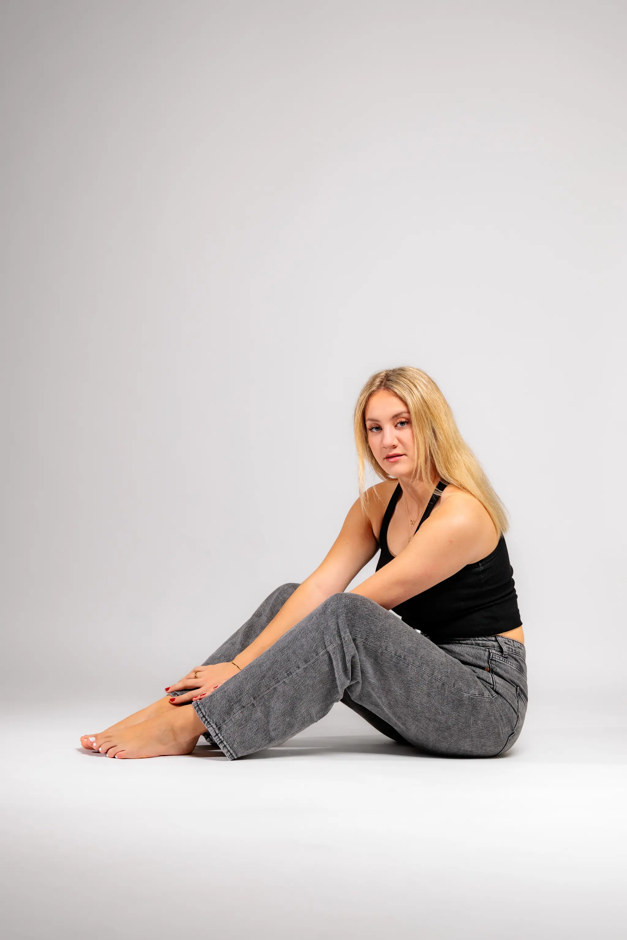 Model Anna sitzt in einem lässigen Outfit mit schwarzem Top und grauer Jeans barfuß auf dem weißen Studioboden und blickt in die Kamera