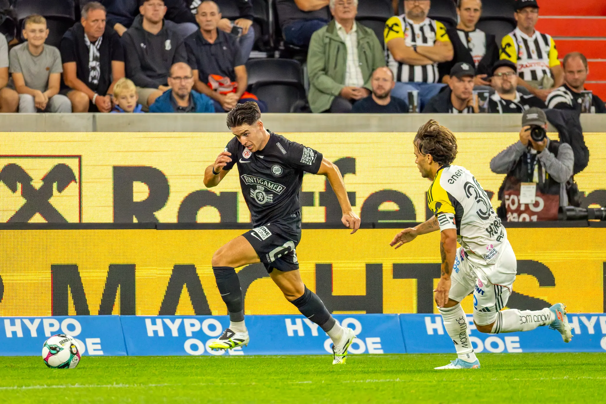 Ein Fußballspieler im schwarzen Trikot rennt dynamisch mit dem Ball auf dem Feld, verfolgt von einem Gegenspieler in hellem Trikot, mit einer großen LED-Werbewand und Zuschauern im Hintergrund