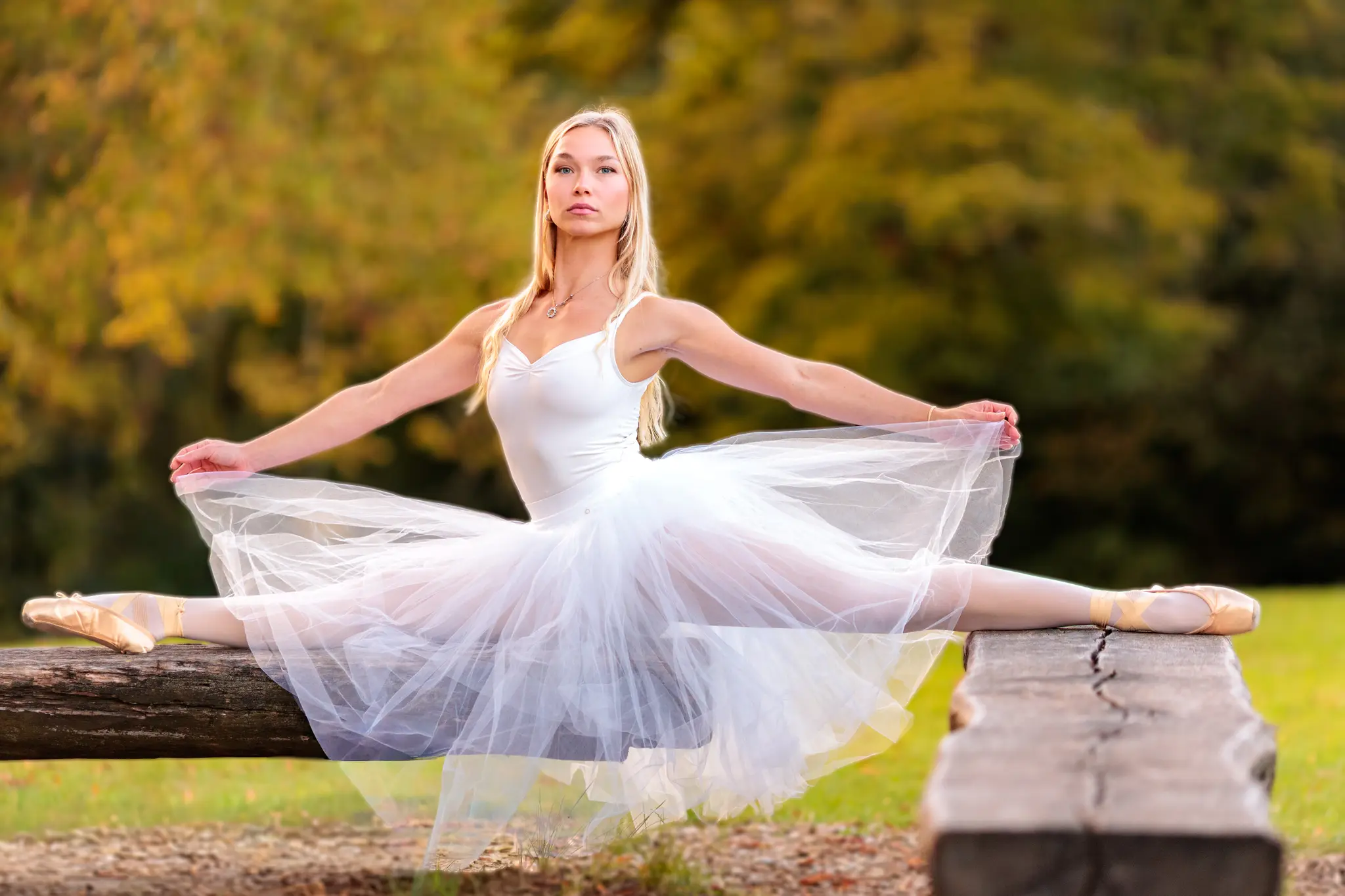 Eine anmutige Balletttänzerin in einem weißen Tutu vollführt einen Spagat auf einem rustikalen Holzbalken in der Natur