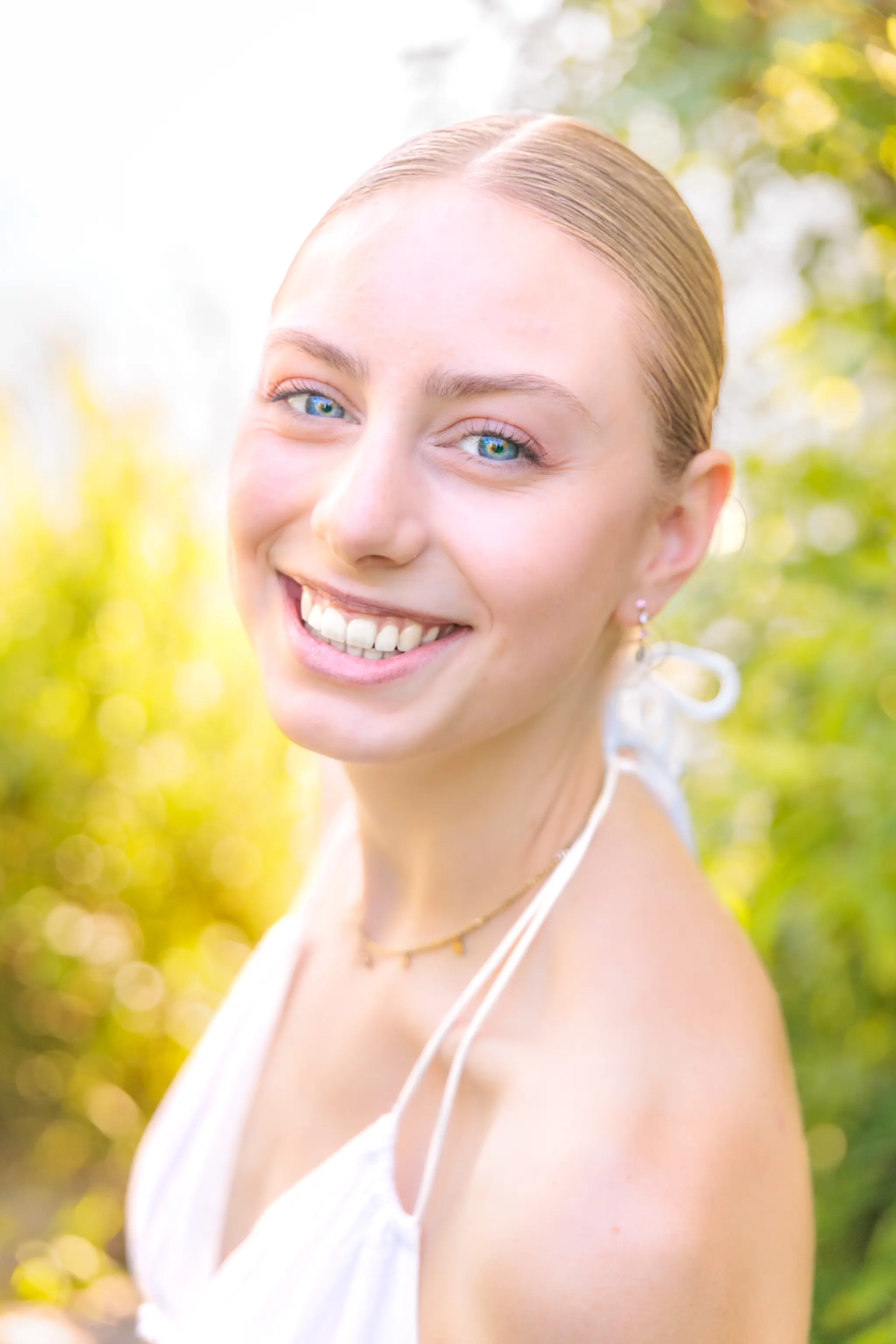 Porträt einer lächelnden jungen Frau mit blonden Haaren, blauen Augen und einem weißen Kleid im Freien mit verschwommenem Hintergrund aus grünen Blättern und goldenem Licht. 