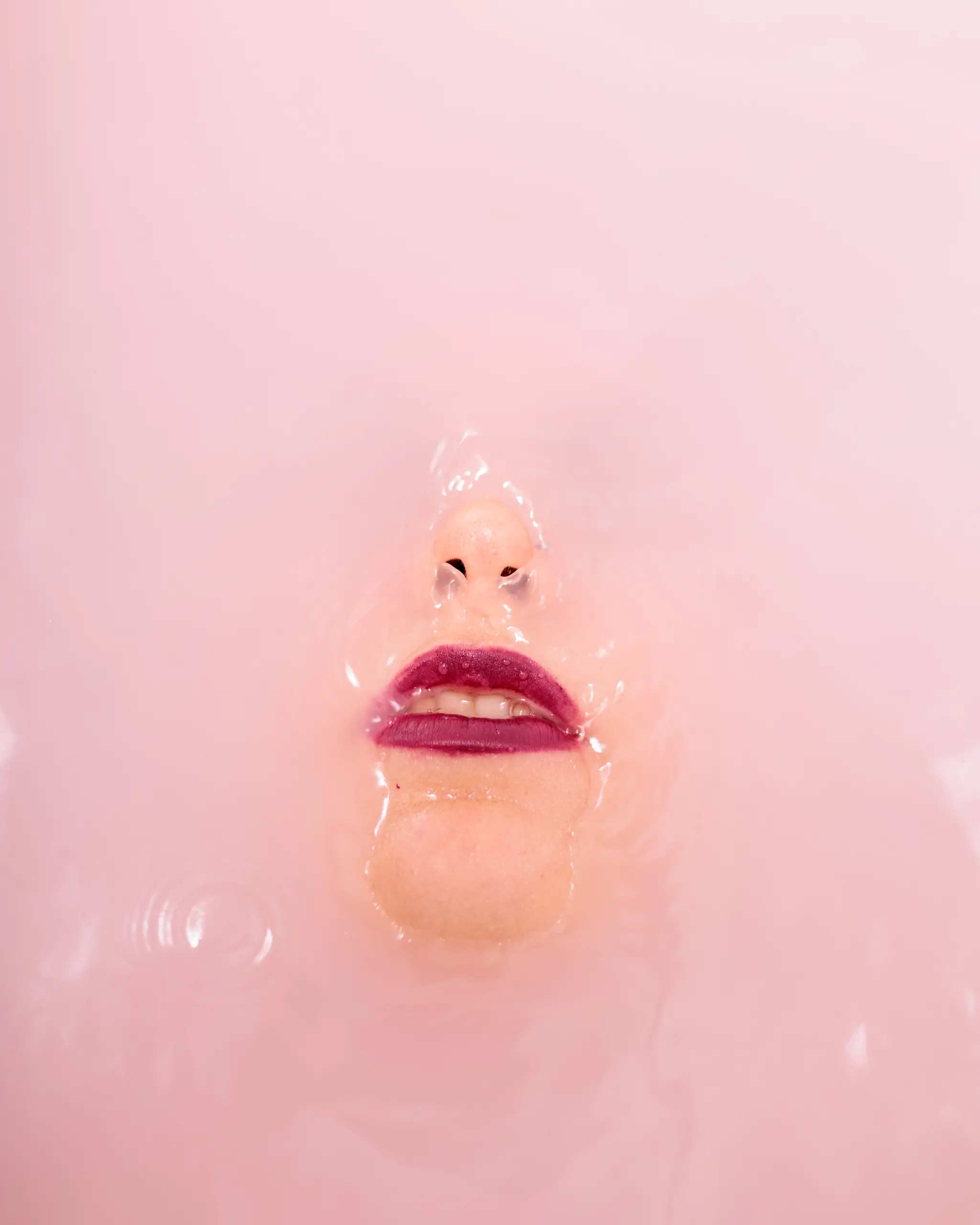 Ein Gesicht mit lila Lippenstift ist teilweise in einem sanften rosa Milchbad zu sehen 