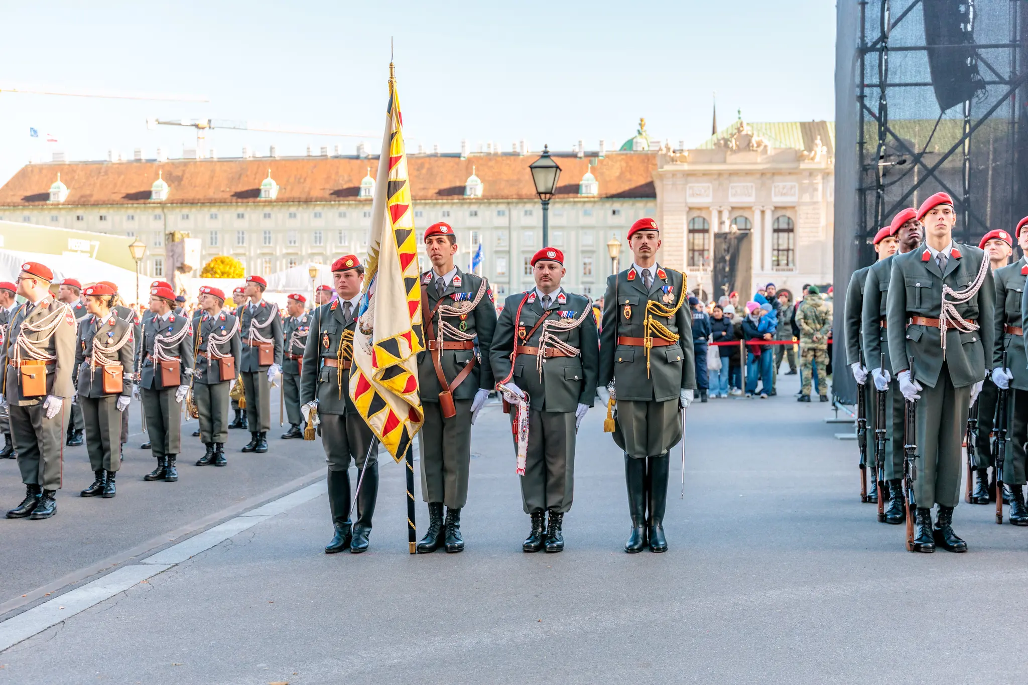 Gardesoldaten in dunkelgrünen Uniformen mit roten Baretten stehen mit einer Fahne in Reih und Glied auf dem Heldenplatz, dahinter die Neue Burg.
