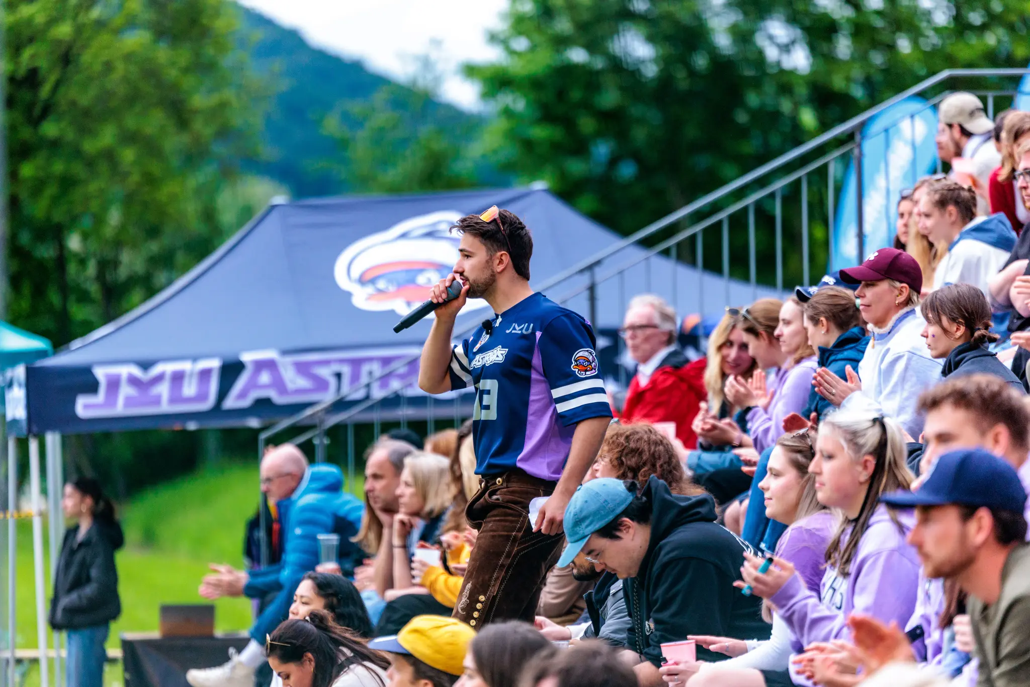 Ein Mann in Sporttrikot und Lederhose spricht bei einem Sportevent ins Mikrofon, während Zuschauer applaudieren 