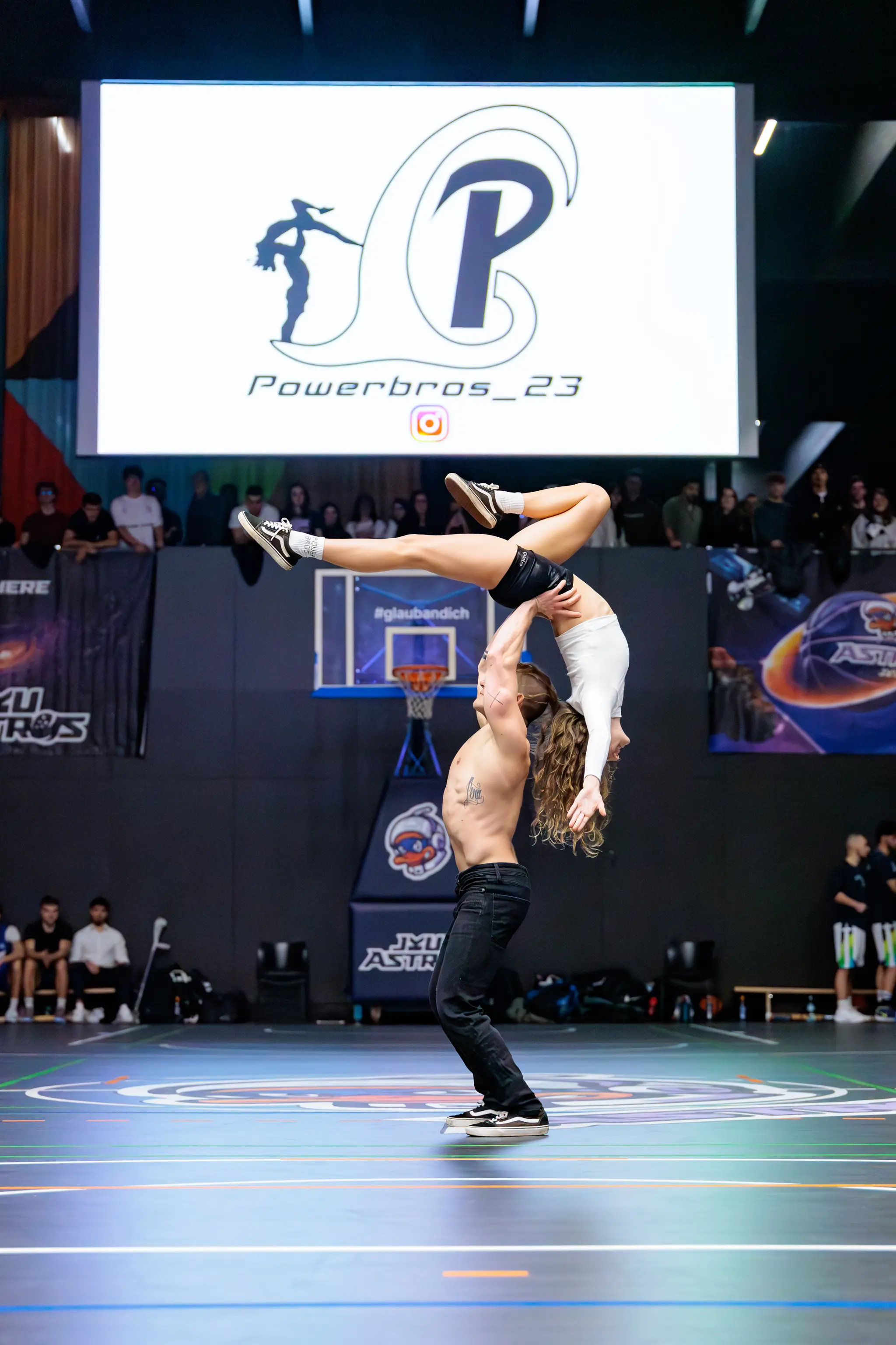 Eine Turnvorführung präsentiert einen Mann, der eine Frau in einer eleganten Hebefigur in einer Sporthalle mit großem "Powerbros" Bildschirm trägt