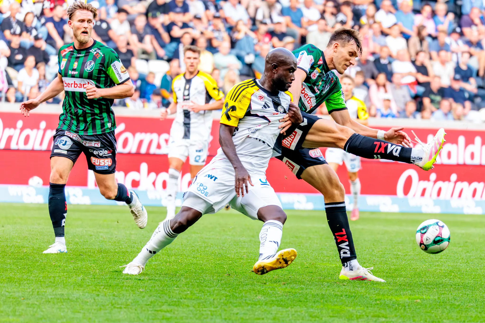 Zwei Fußballspieler, einer vom LASK im grün-schwarzen Trikot und ein Spieler von SV Ried im weiß-gelben Trikot, kämpfen in einem dynamischen Zweikampf um den Ball während eines Bundesligaspiels