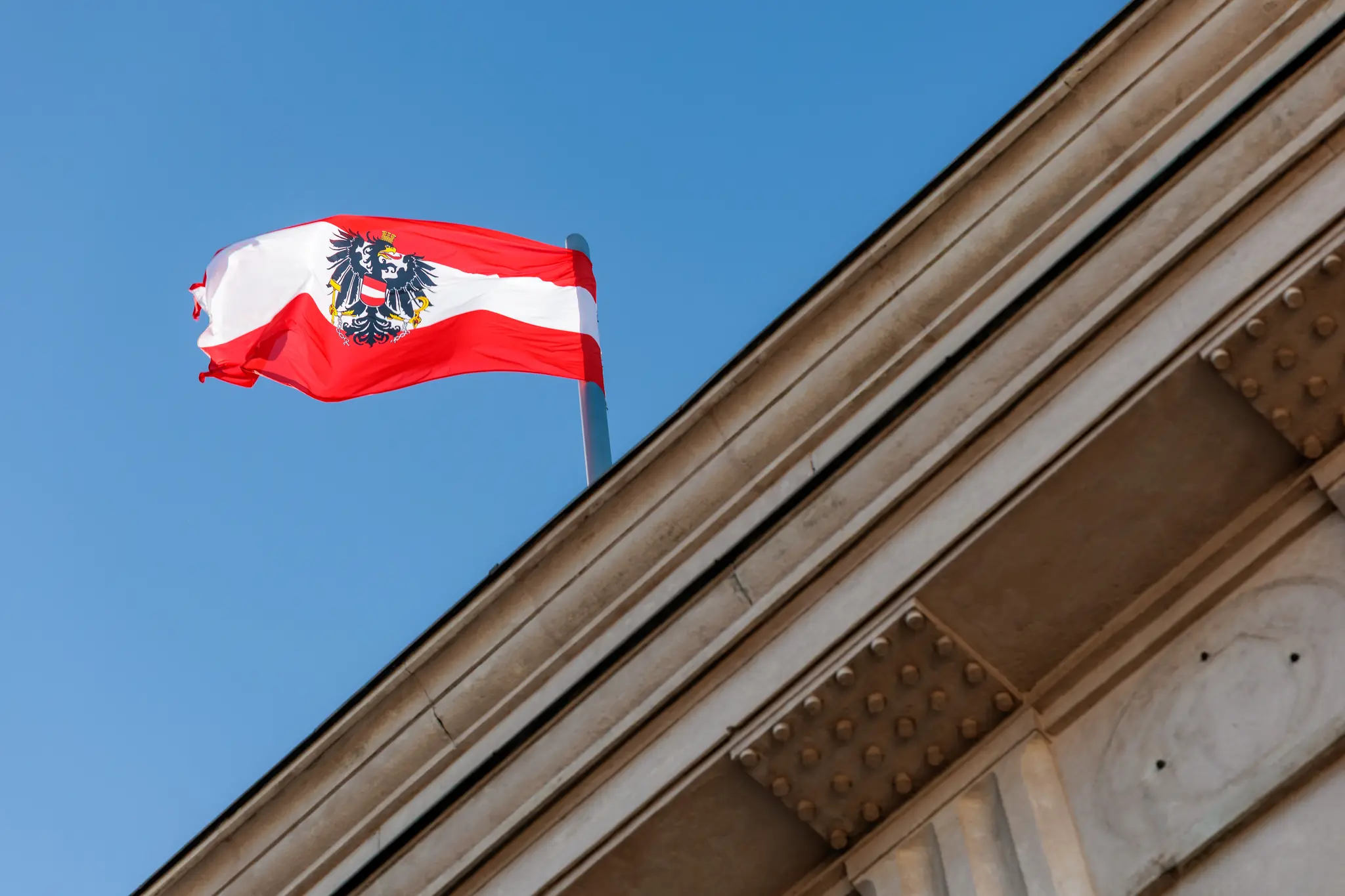 Die österreichische Flagge mit Bundesadler weht im Wind über dem Dach der Hofburg, dahinter ein tiefblauer Himmel.