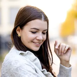 Eine lächelnde junge Frau mit dunklem Haar und hellem Strickpullover trägt einen herzförmigen Ohrring und blickt nach unten lens: 56mm F1.4 DC DN | Contemporary 018