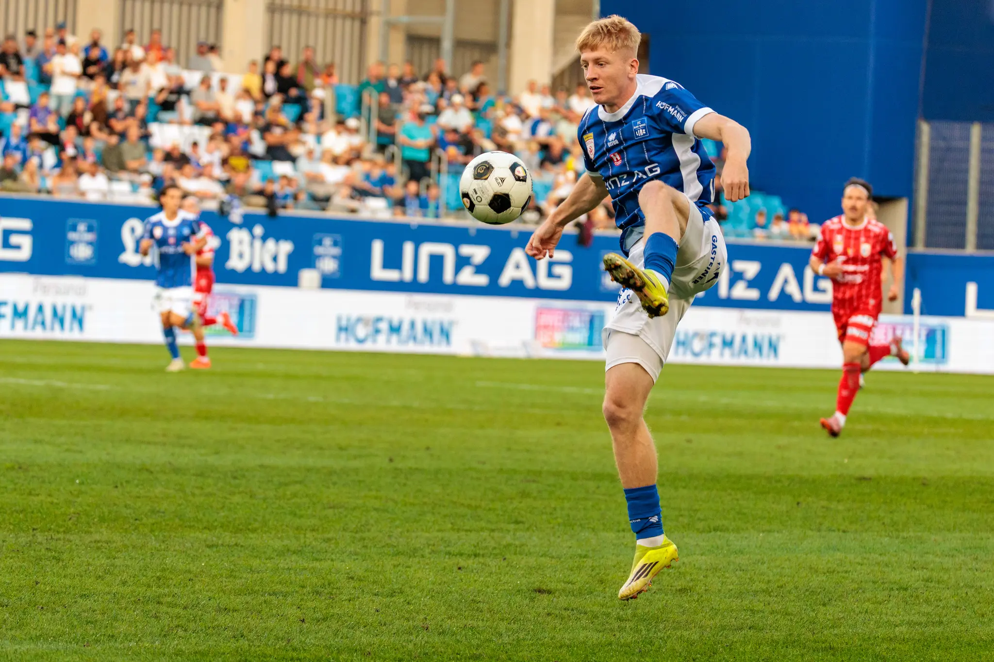 Ein Linzer Spieler nimmt einen hohen Ball mit dem Oberschenkel an; im Hintergrund läuft ein Welser Spieler mit Kopfband, daneben ist die Linz-AG-Bande zu sehen.