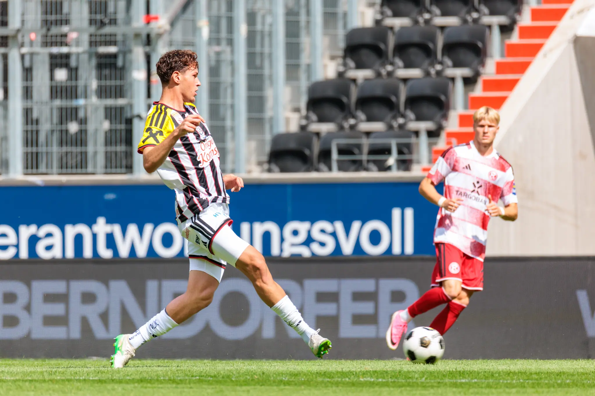 LASK-Spieler im schwarz-weiß-gelben Trikot kontrolliert den Ball am Fuß und dribbelt nach vorne, während ein Düsseldorfer Spieler herannaht.