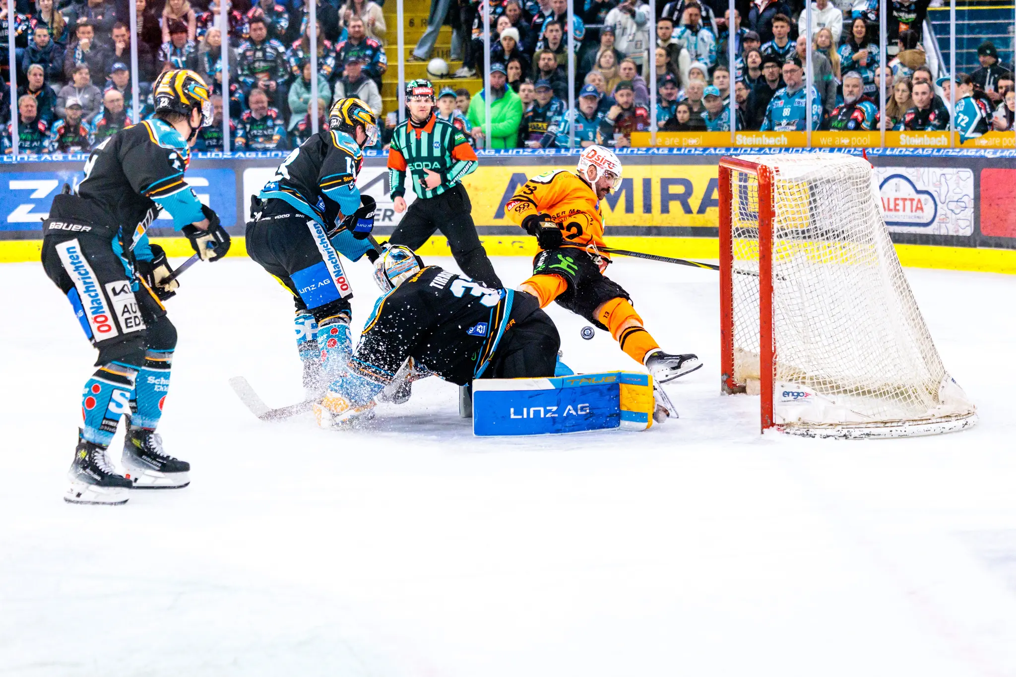 Eishockey-Spielszene vor dem Tor, bei der ein Spieler in orangefarbenem Trikot gegen den am Boden liegenden Torhüter und einen Verteidiger in schwarz-türkisem Dress im Kampf um den Puck ist 