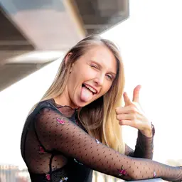 Eine fröhliche blonde Frau in einem transparenten Oberteil zwinkert, streckt die Zunge heraus und zeigt einen Daumen nach oben lens: 56mm F1.4 DC DN | Contemporary 018
