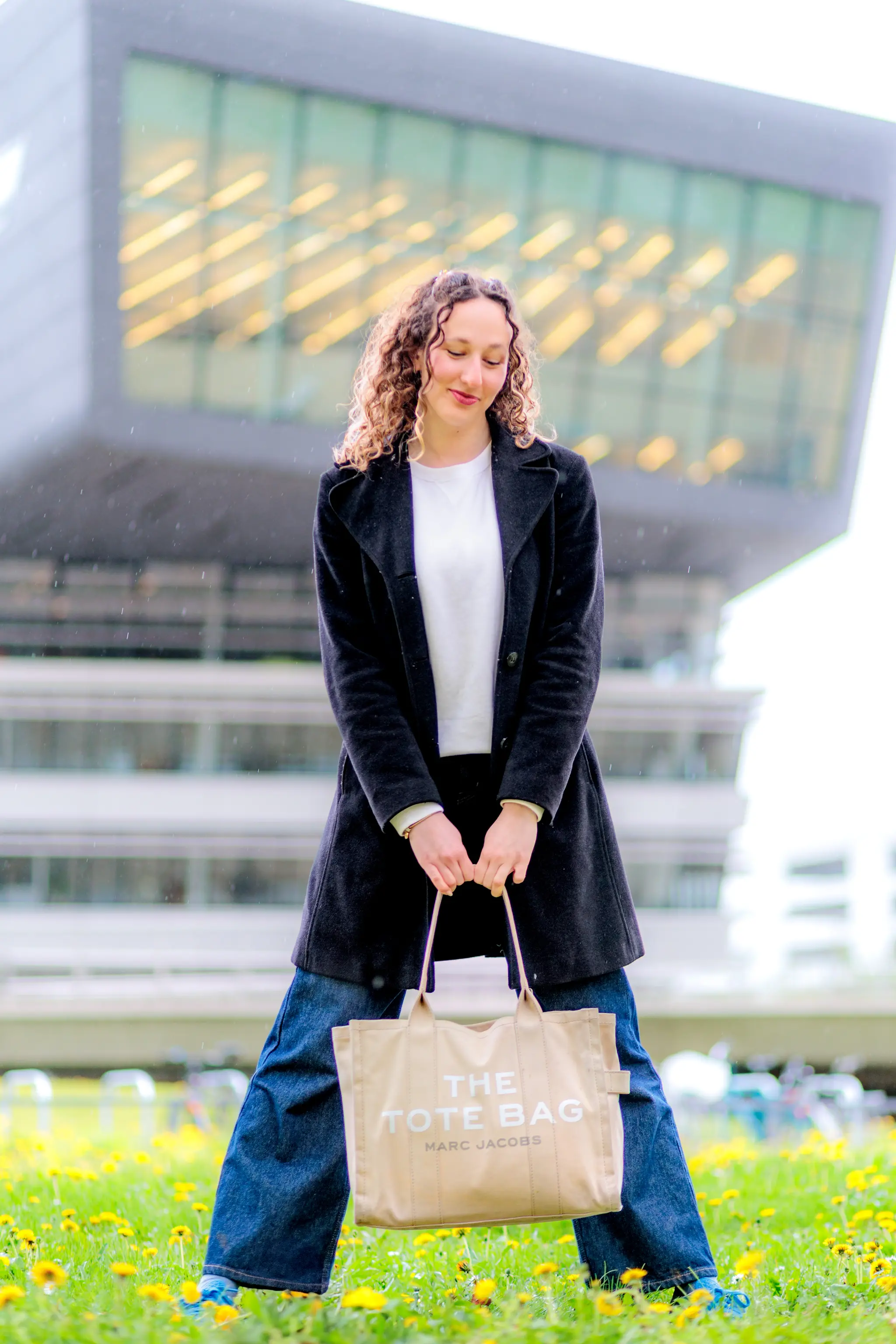 Junge Frau in schwarzem Mantel und Jeans hält eine beige Marc Jacobs Tote Bag vor einem modernen Gebäude auf einer blumenübersäten Wiese 