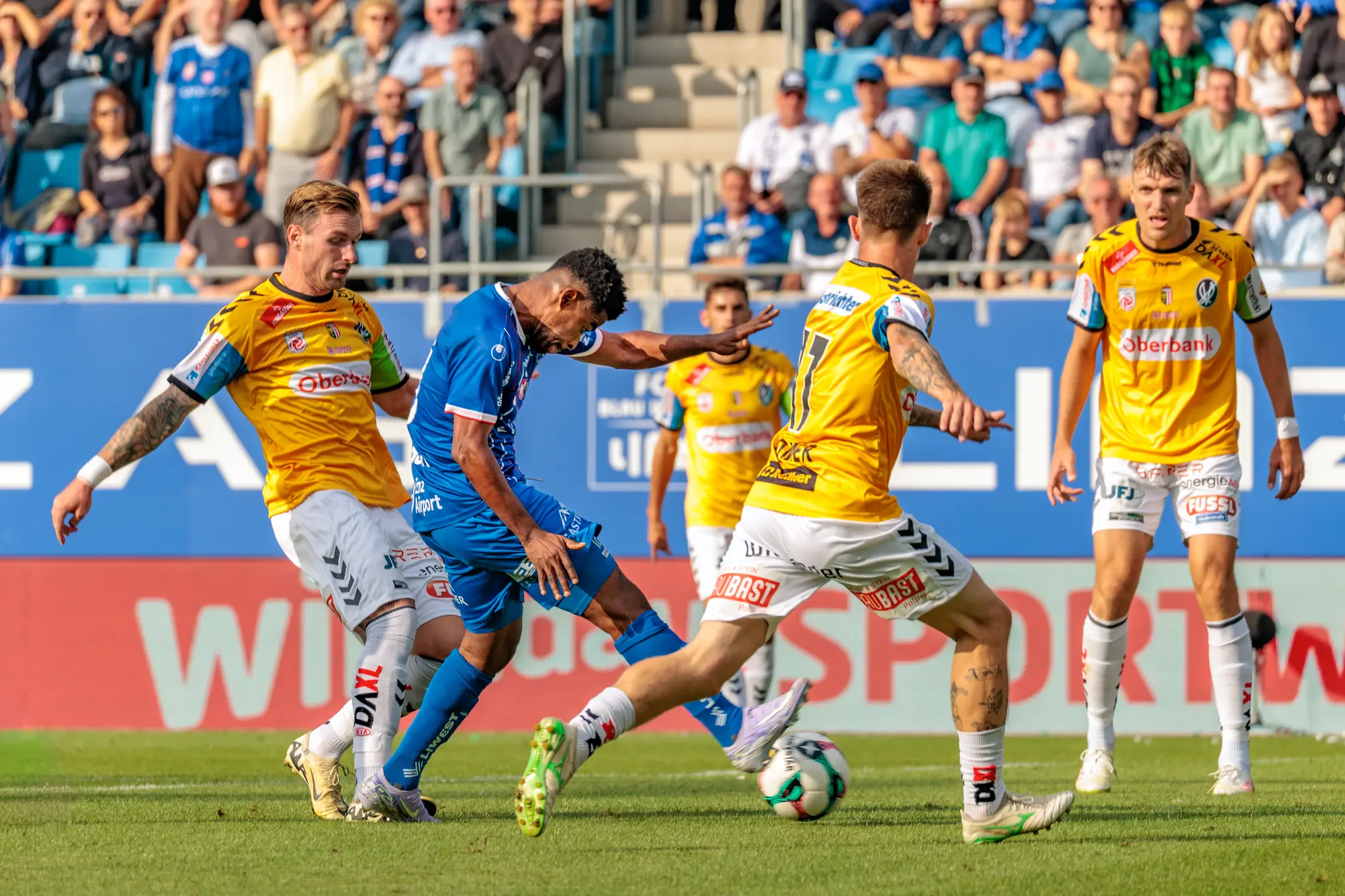 Intensiver Zweikampf um den Ball zwischen Fußballspielern auf dem Spielfeld, beobachtet von der Zuschauerkulisse im Stadion