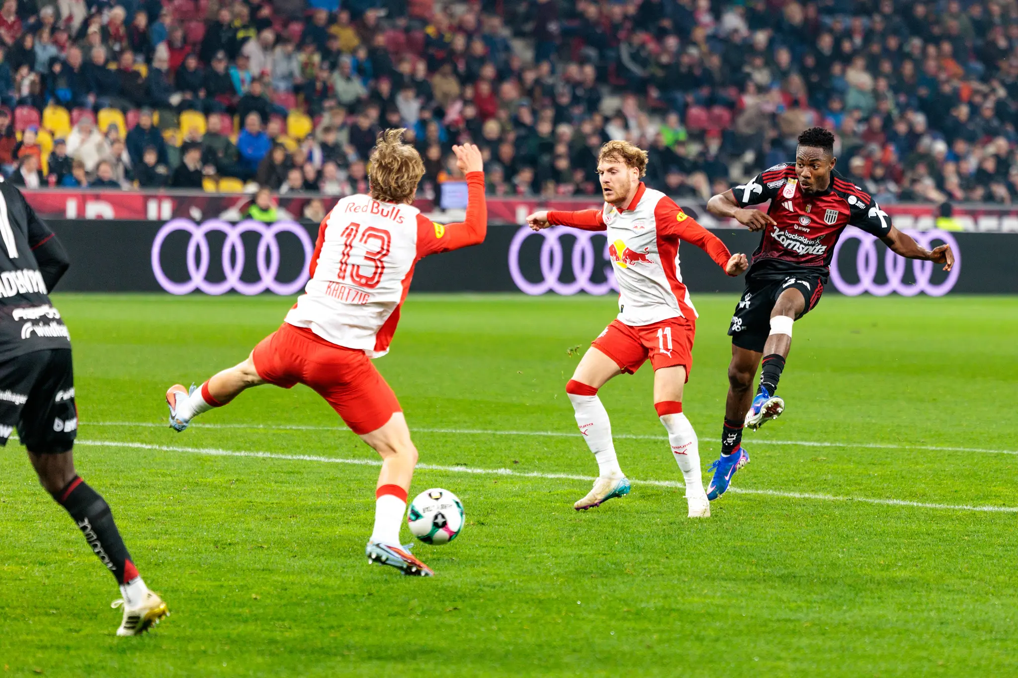 Ein LASK-Angreifer schießt den Ball in Richtung des Salzburger Tores, während zwei Verteidiger von Red Bull Salzburg versuchen, den Schuss in einer intensiven Zweikampfsituation zu blocken