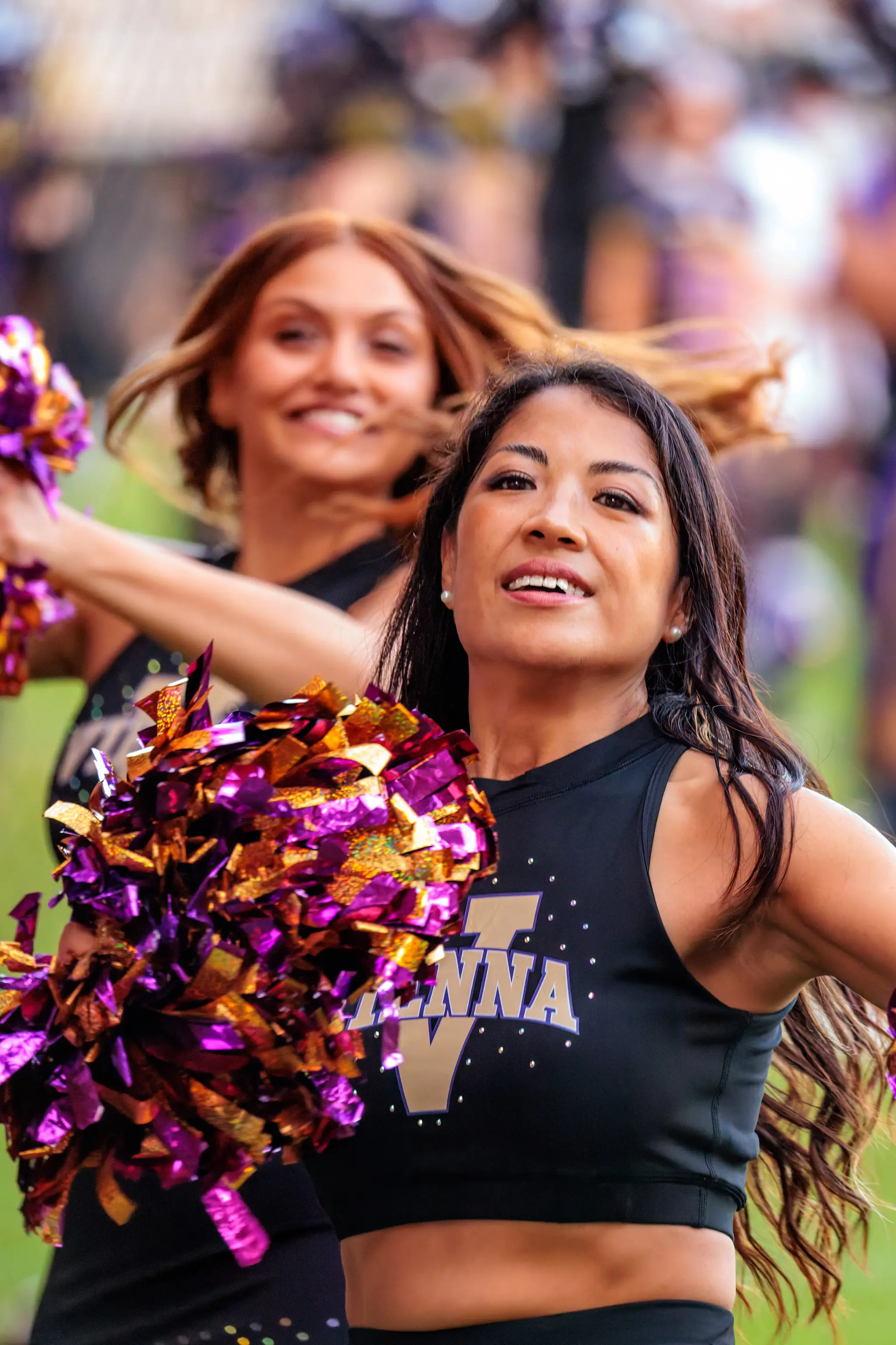 Zwei Tänzerinnen des Vienna Vikings Cheer Squads mitten im Tanz: lachend und im Schritt, die lila-goldenen Pompons in Bewegung.