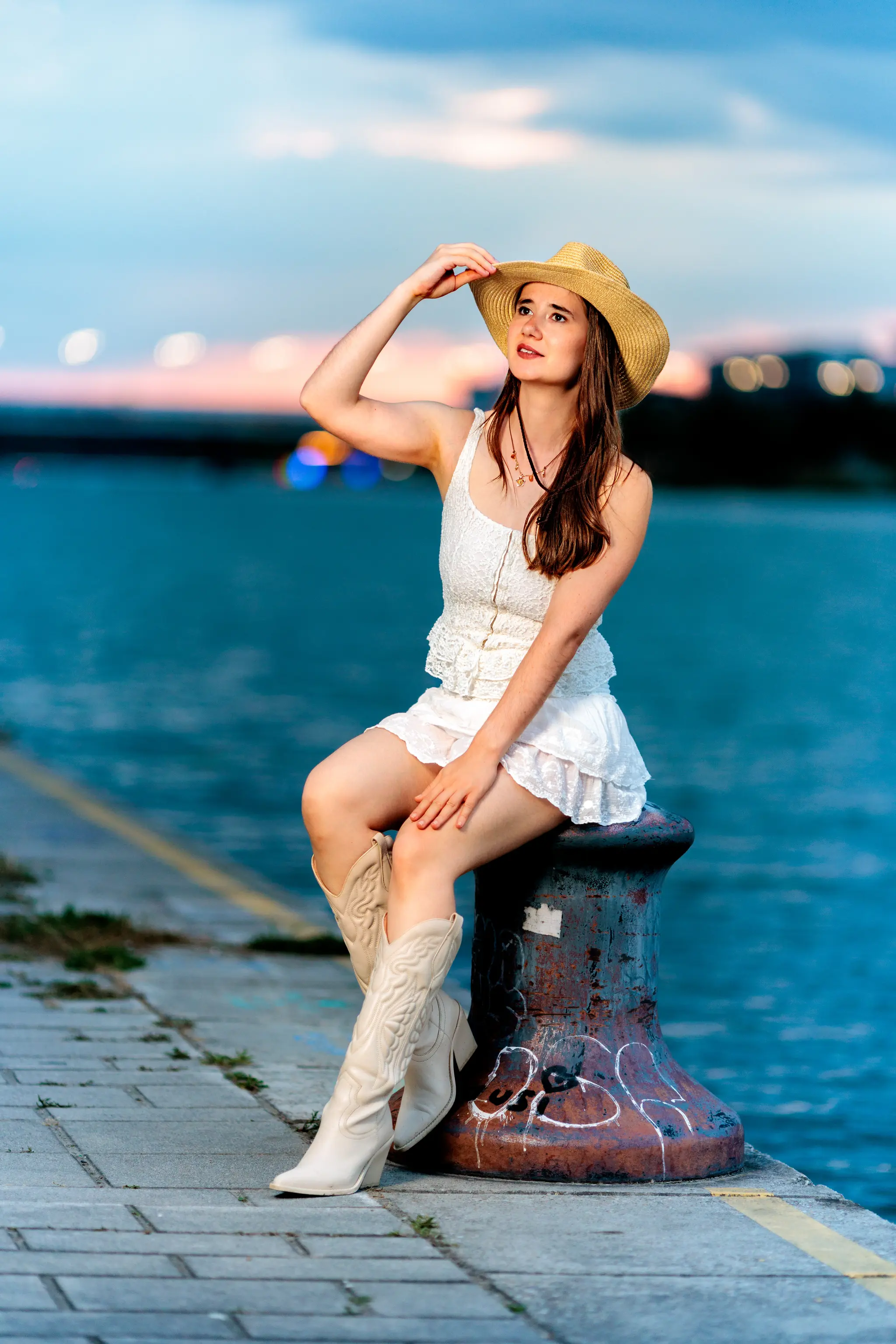 Hanna sitzt am Wasser auf einem Bollwerk, berührt den Rand ihres Strohhuts und schaut in der Dämmerung in die Kamera.