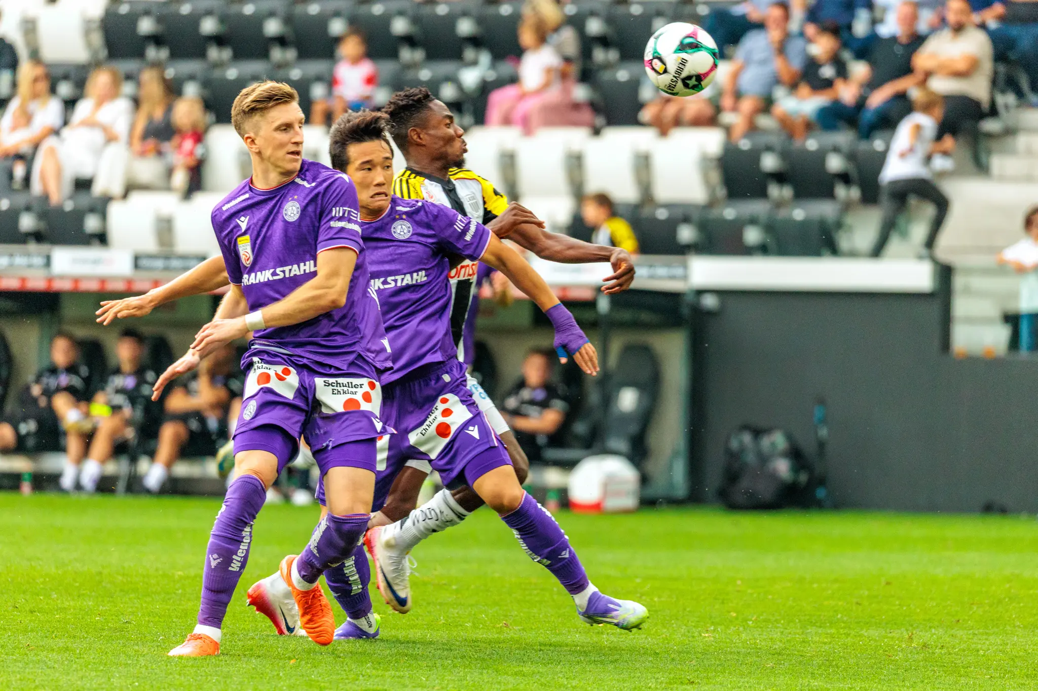 Ein intensiver Zweikampf zwischen drei Fußballspielern, zwei in violetten und einem in einem schwarz-gelben Trikot, um den schwebenden Ball während eines Spiels der Bundesliga