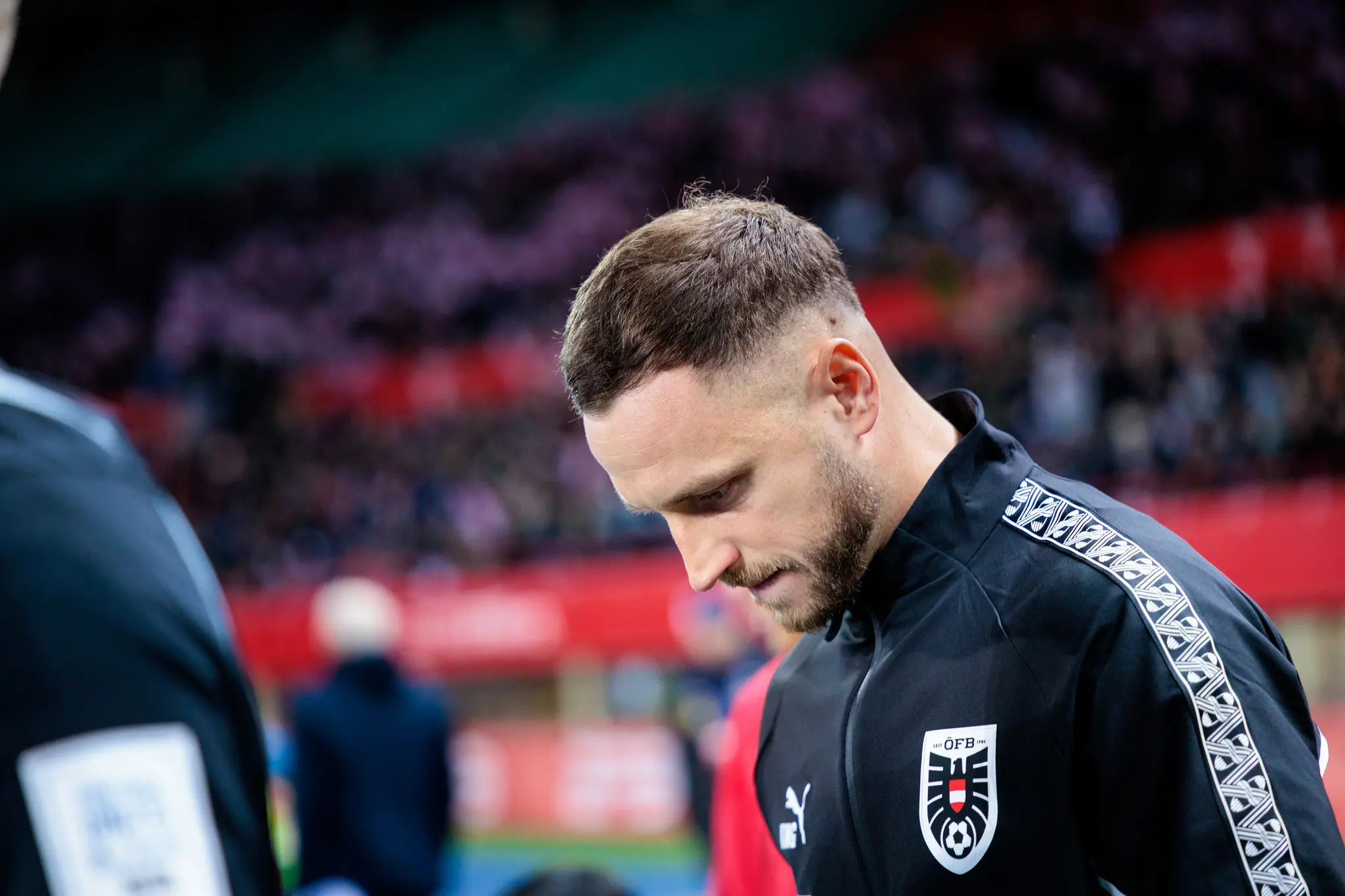 Ein österreichischer Nationalspieler in schwarzer ÖFB-Teamjacke mit markantem Ärmeldesign blickt im Stadion nach unten