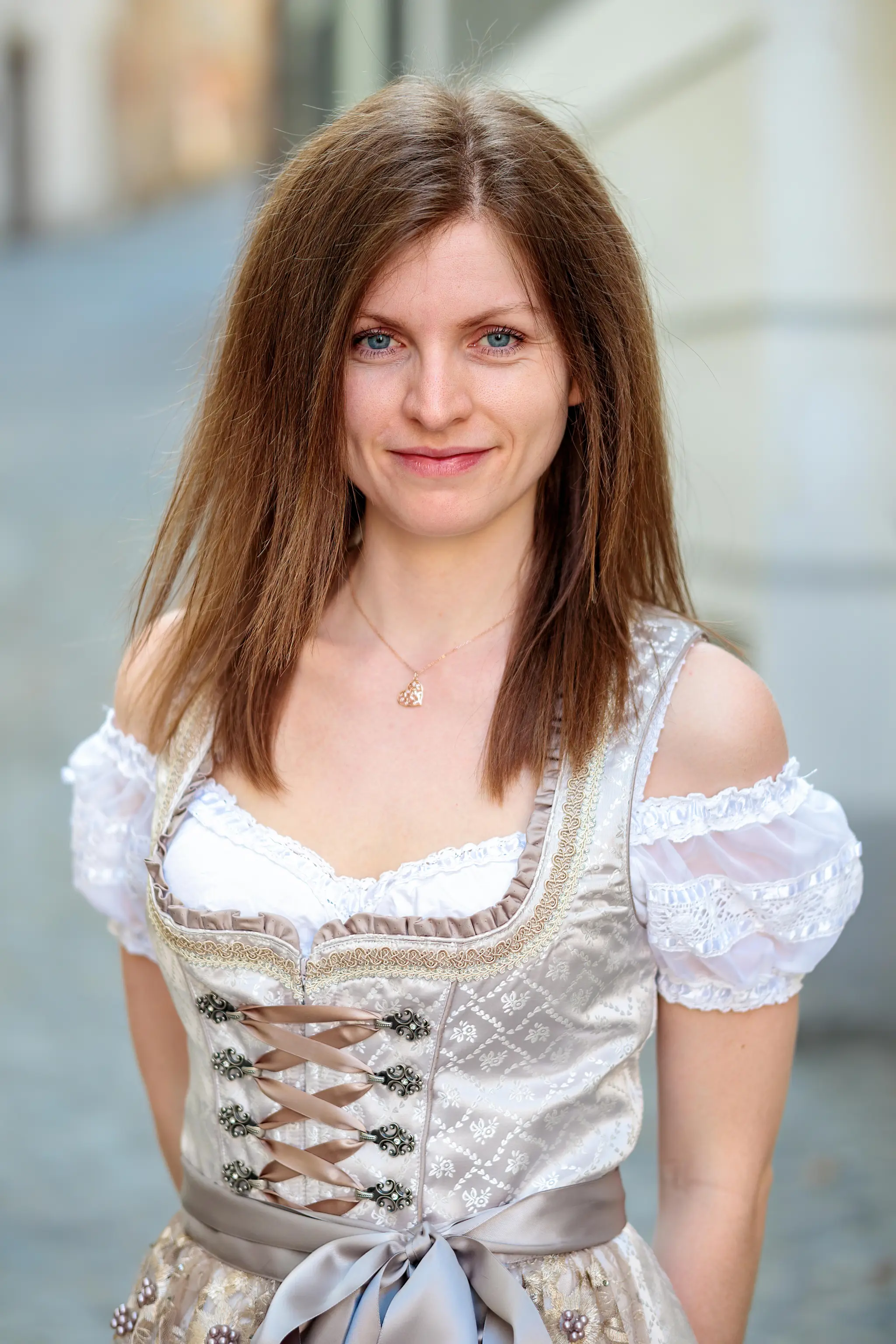 Porträt einer jungen Frau in einem traditionellen hellen Dirndl mit Spitzenbluse und Herz-Halskette, die freundlich in die Kamera lächelt
