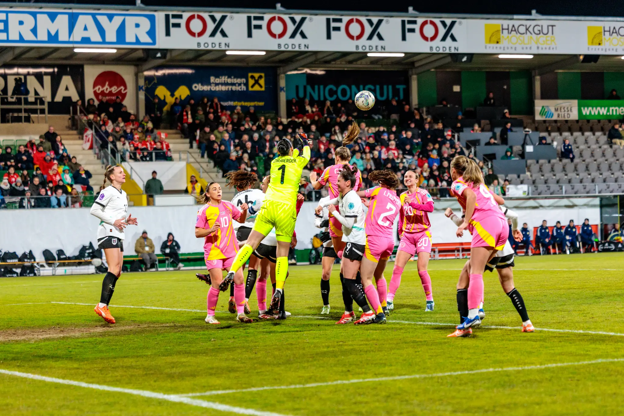 Eine Torhüterin in leuchtend gelbem Trikot springt bei einem Frauenfußballspiel nach dem Ball, umgeben von Spielerinnen in weißen und pinken Trikots und zahlreichen Zuschauern auf den Tribünen 