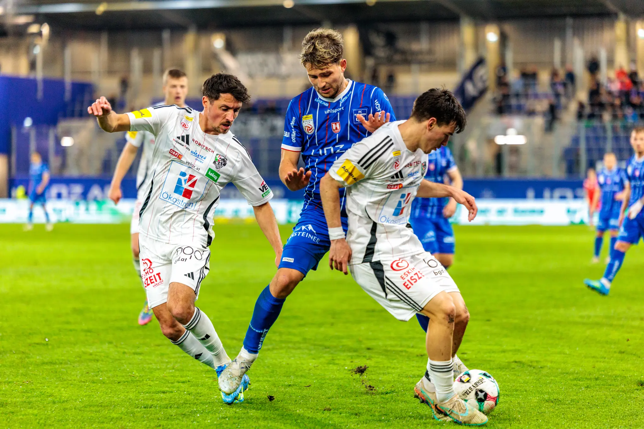 Ein Spieler in blauen Trikot kämpft unter Bedrängnis zweier Gegner in weißen Dress um den Ballbesitz