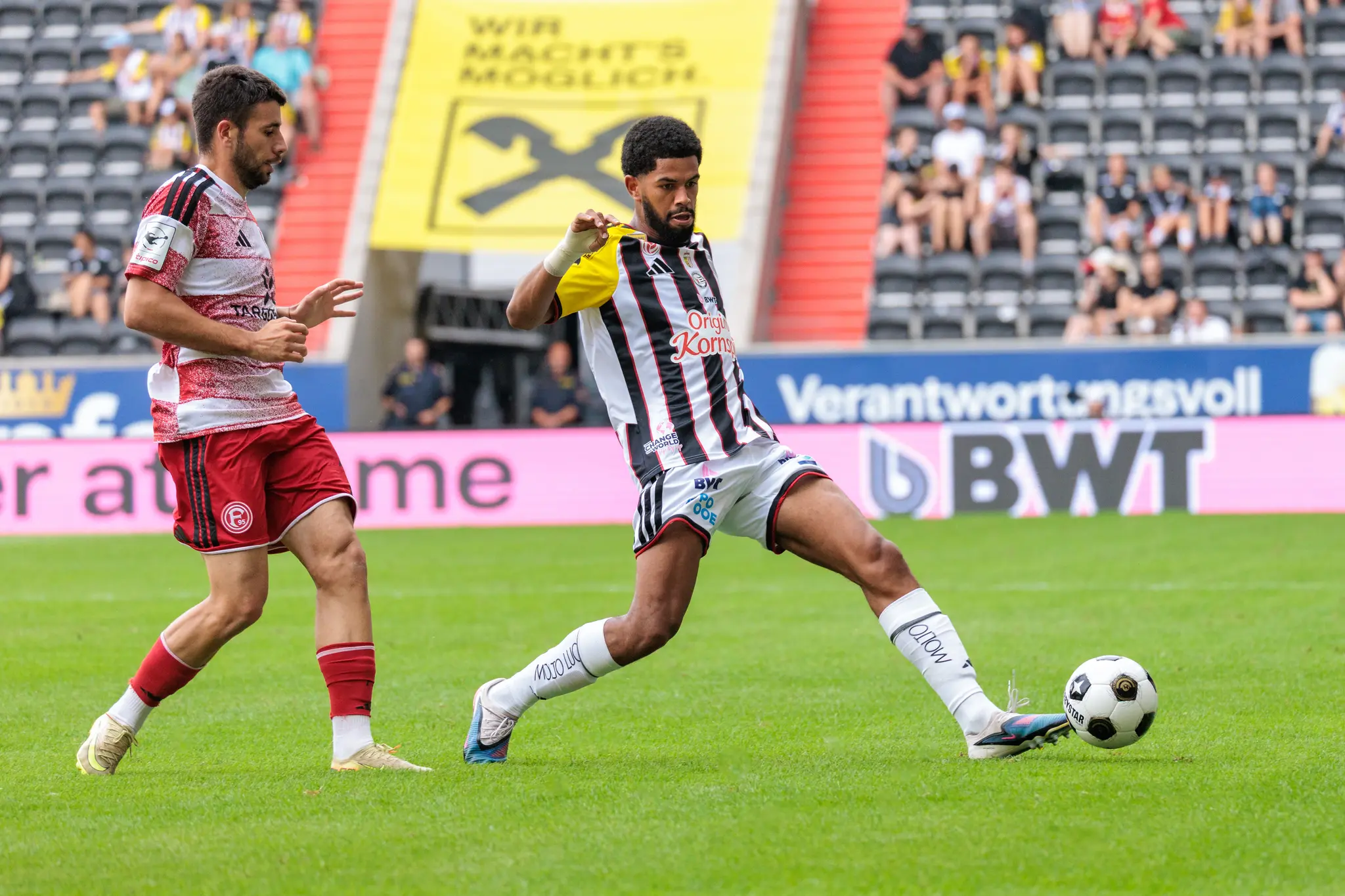 LASK-Spieler kontrolliert den Ball am Boden und bereitet den Spielaufbau vor, im Hintergrund ein Düsseldorfer Spieler und die Zuschauertribüne.