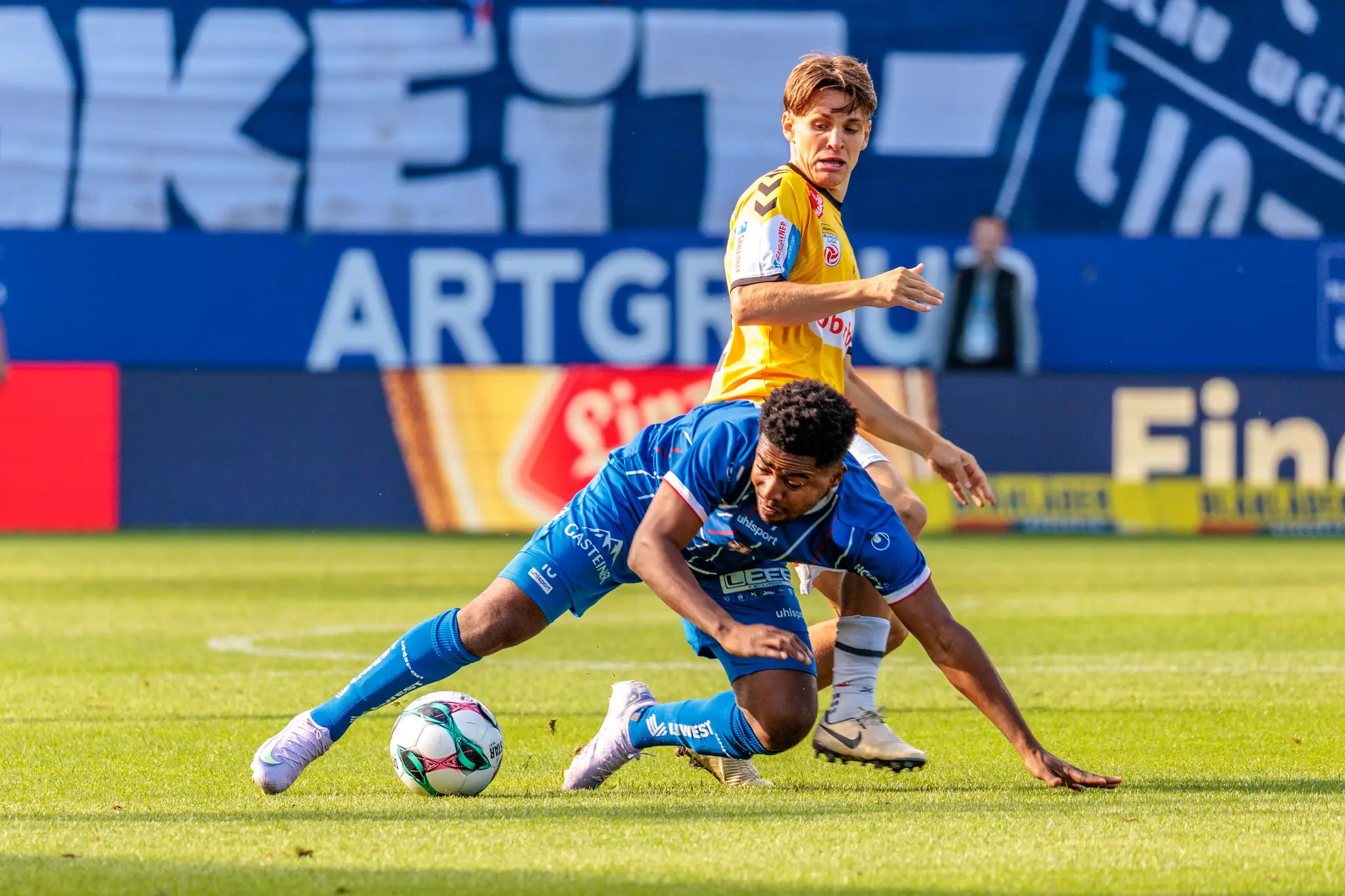 Fußball-Zweikampf im Bundesliga-Derby: Ein Spieler von Blau-Weiß Linz im blauen Trikot geht zu Boden, um den Ball vor einem Gegenspieler von SV Ried im gelben Trikot zu behaupten