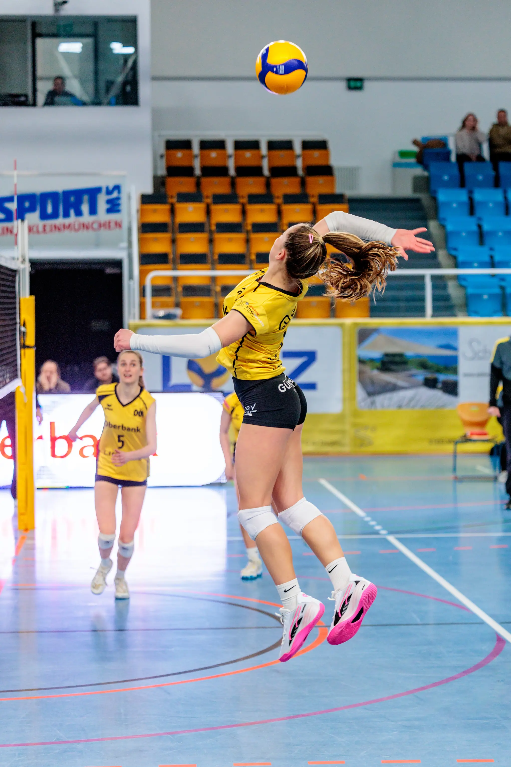 Eine Volleyballspielerin in einem gelben Trikot springt in einer Sporthalle, um einen gelb-blauen Volleyball während eines Spiels zu schlagen 