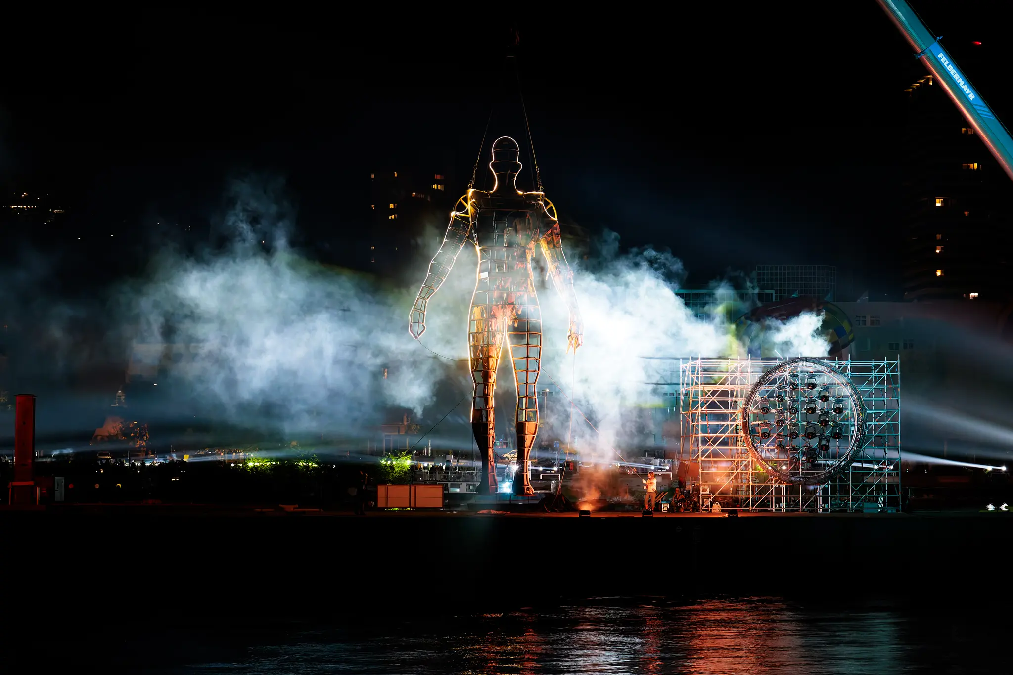 Nächtliche Lichtkunstinstallation mit einer riesigen, goldleuchtenden Drahtgitter-Menschenfigur in Rauch und Nebel an einem Hafen. Eine große runde 2024/09/06