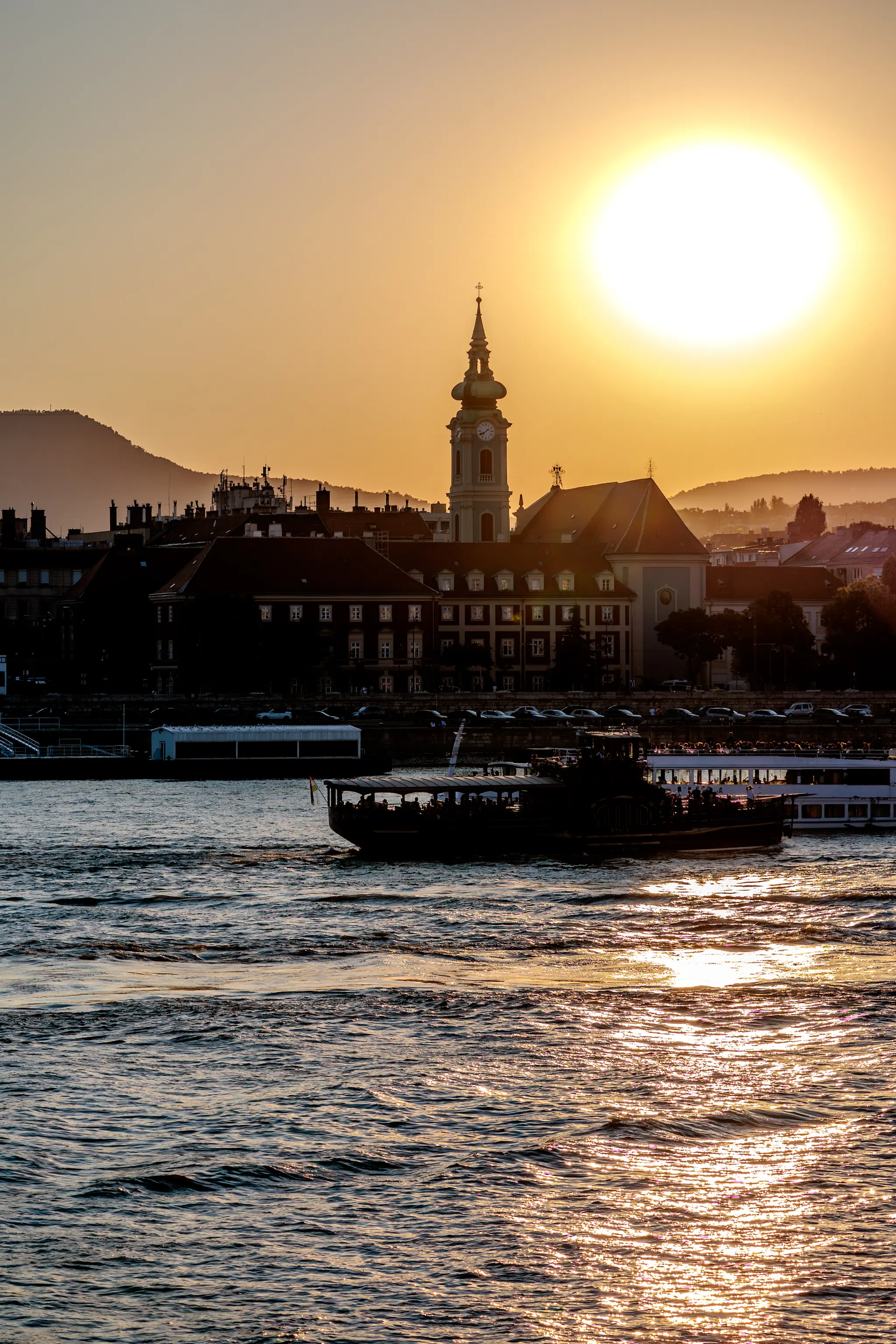 Goldener Sonnenuntergang über der Budapester Skyline mit St. Anna-Kirche, Gebäuden und Booten auf der glitzernden Donau. 