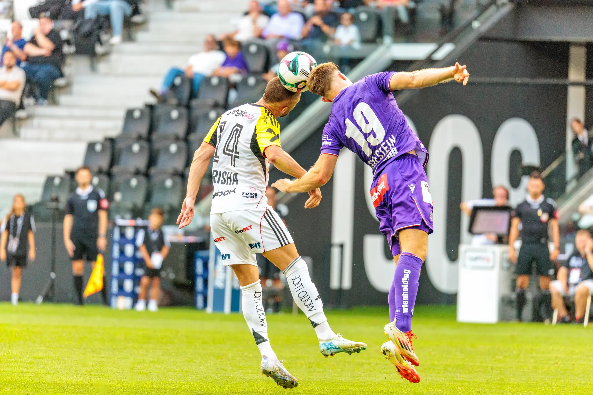 Zwei Fußballspieler in schwarz-weißen und violetten Trikots springen für ein Kopfballduell in die Luft