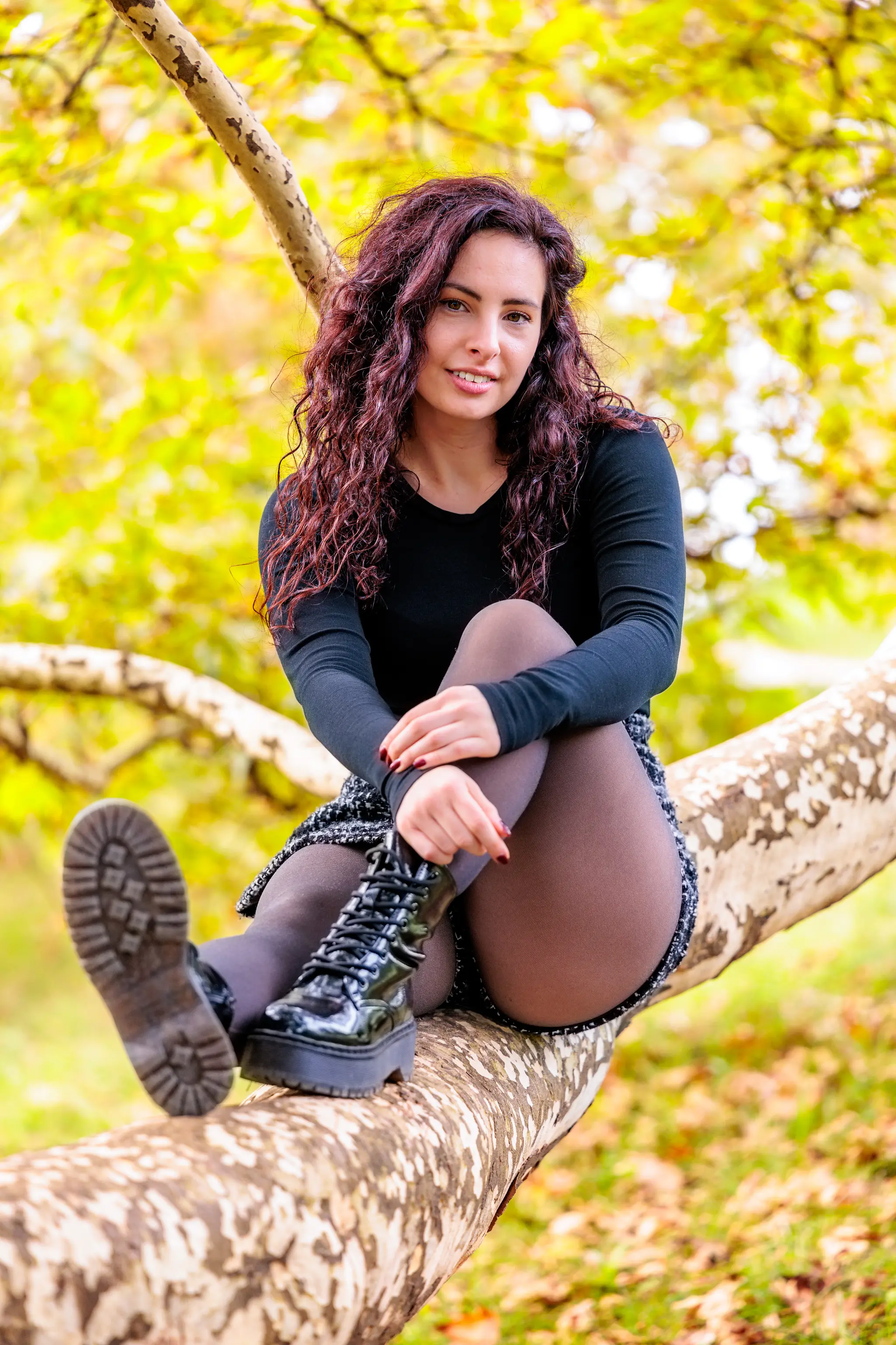 Porträt einer lächelnden jungen Frau mit dunklen Locken, die im Herbst auf einem Baumast vor leuchtend gelben Blättern sitzt