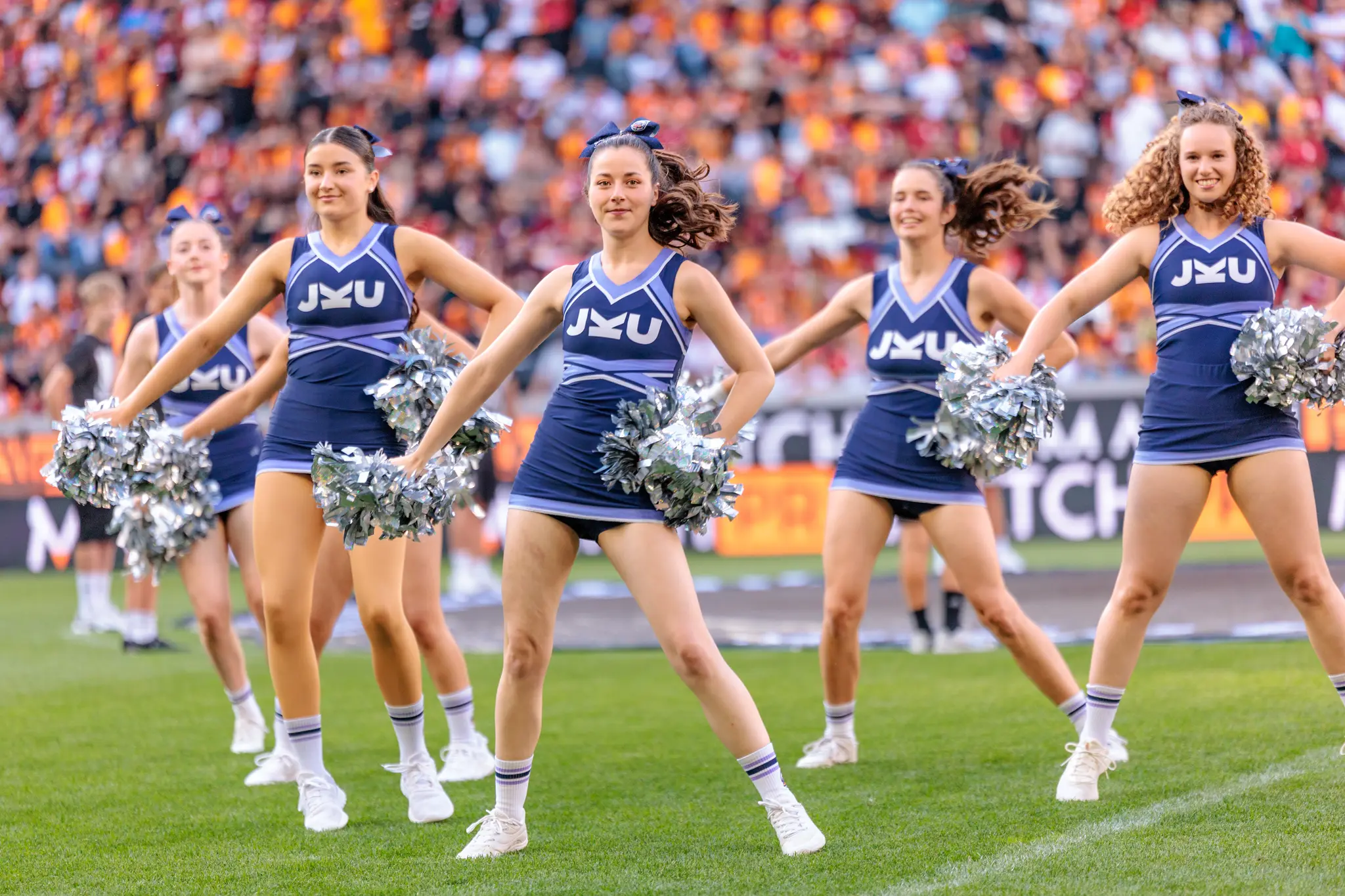 Eine Gruppe junger Cheerleaderinnen in blauen JKU-Uniformen mit silbernen Pompons tritt vor einer Stadionkulisse auf 