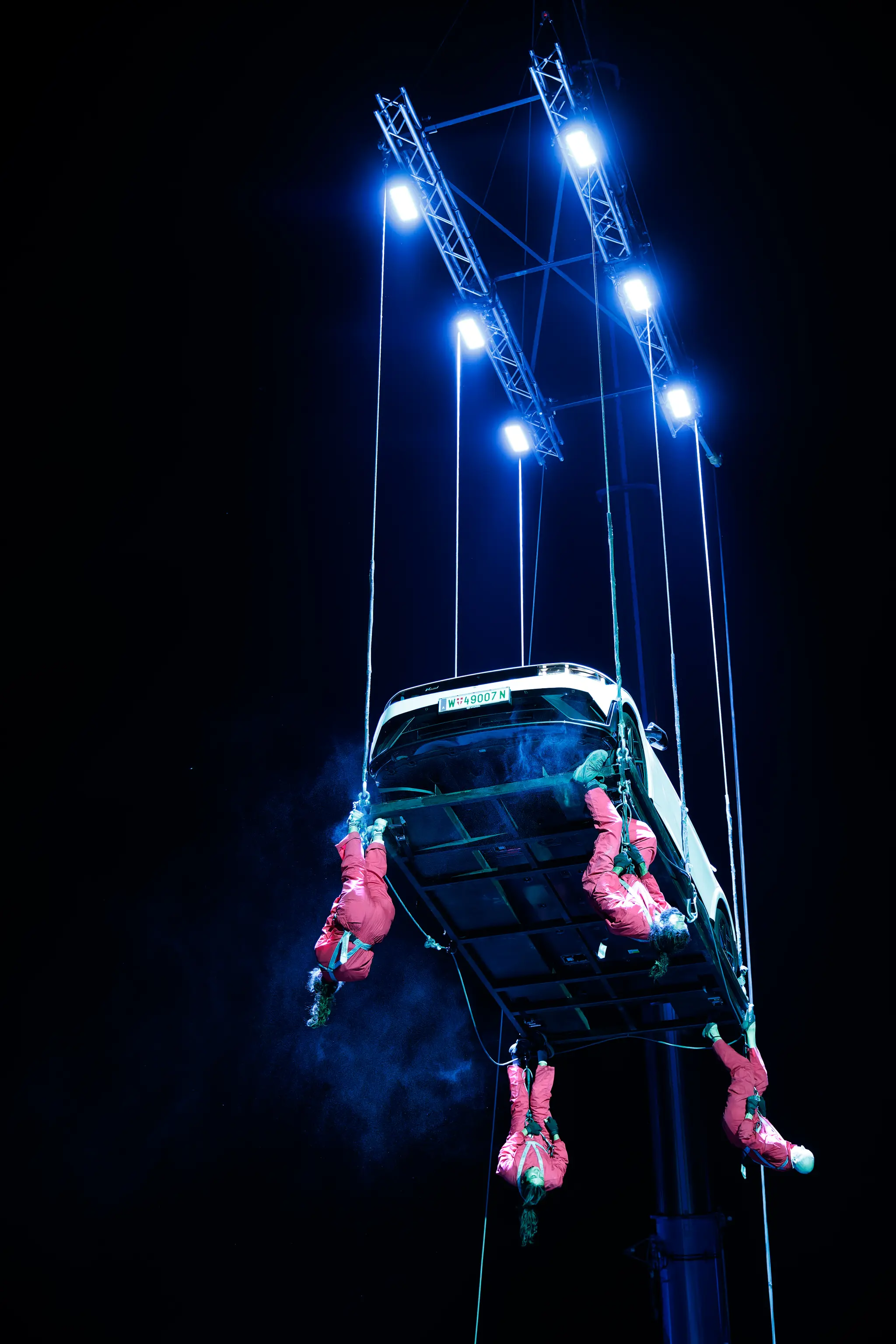 Nächtliche Performance während der Linzer Klangwolke 2024: Ein weißes Auto schwebt an einer blau beleuchteten Traverse, darunter hängen mehrere Personen in roten Overalls kopfüber 