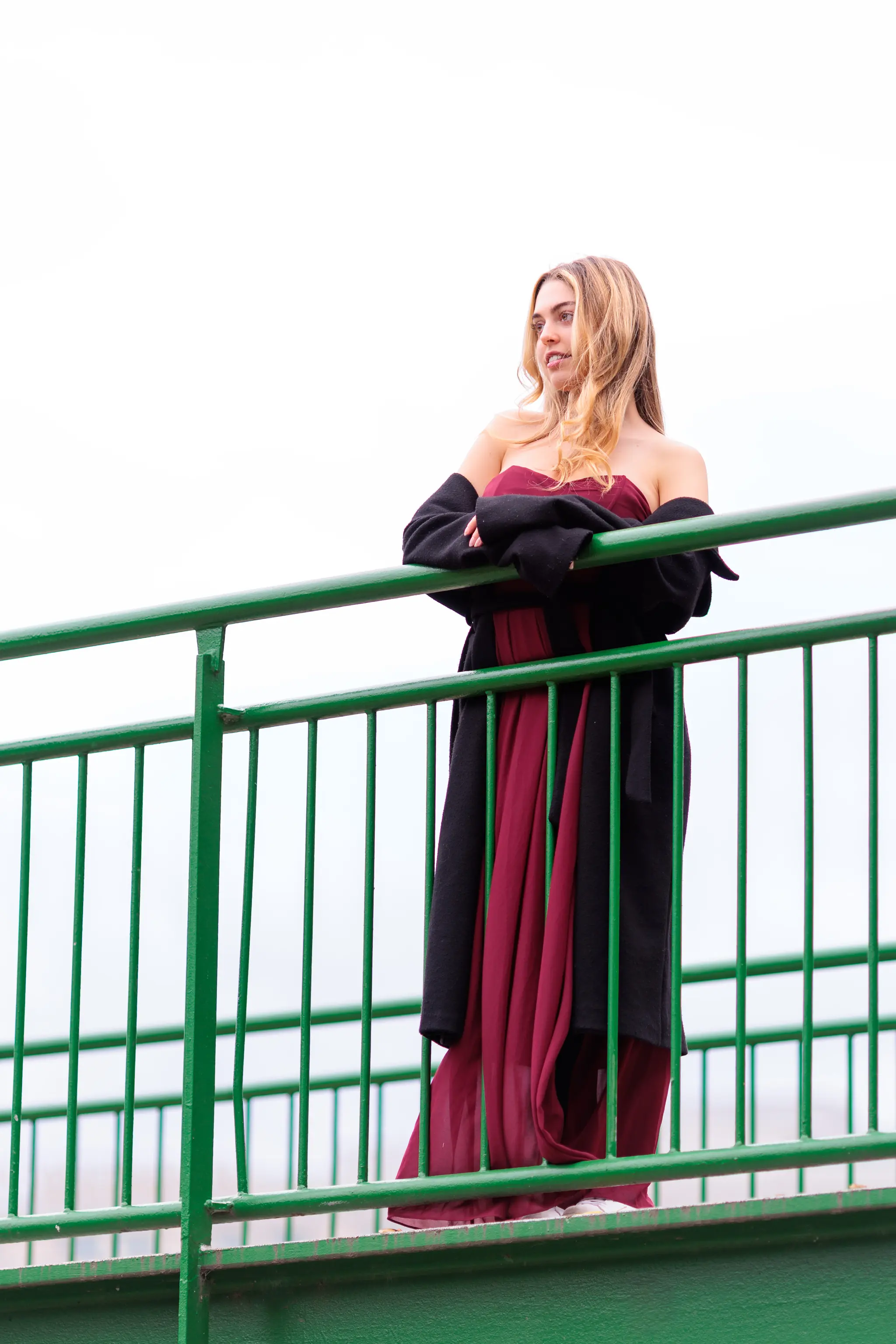 Das Model Valentina lehnt in einem eleganten roten Kleid und einem schwarzen Mantel lächelnd am grünen Geländer einer Brücke im Wiener Stadtpark