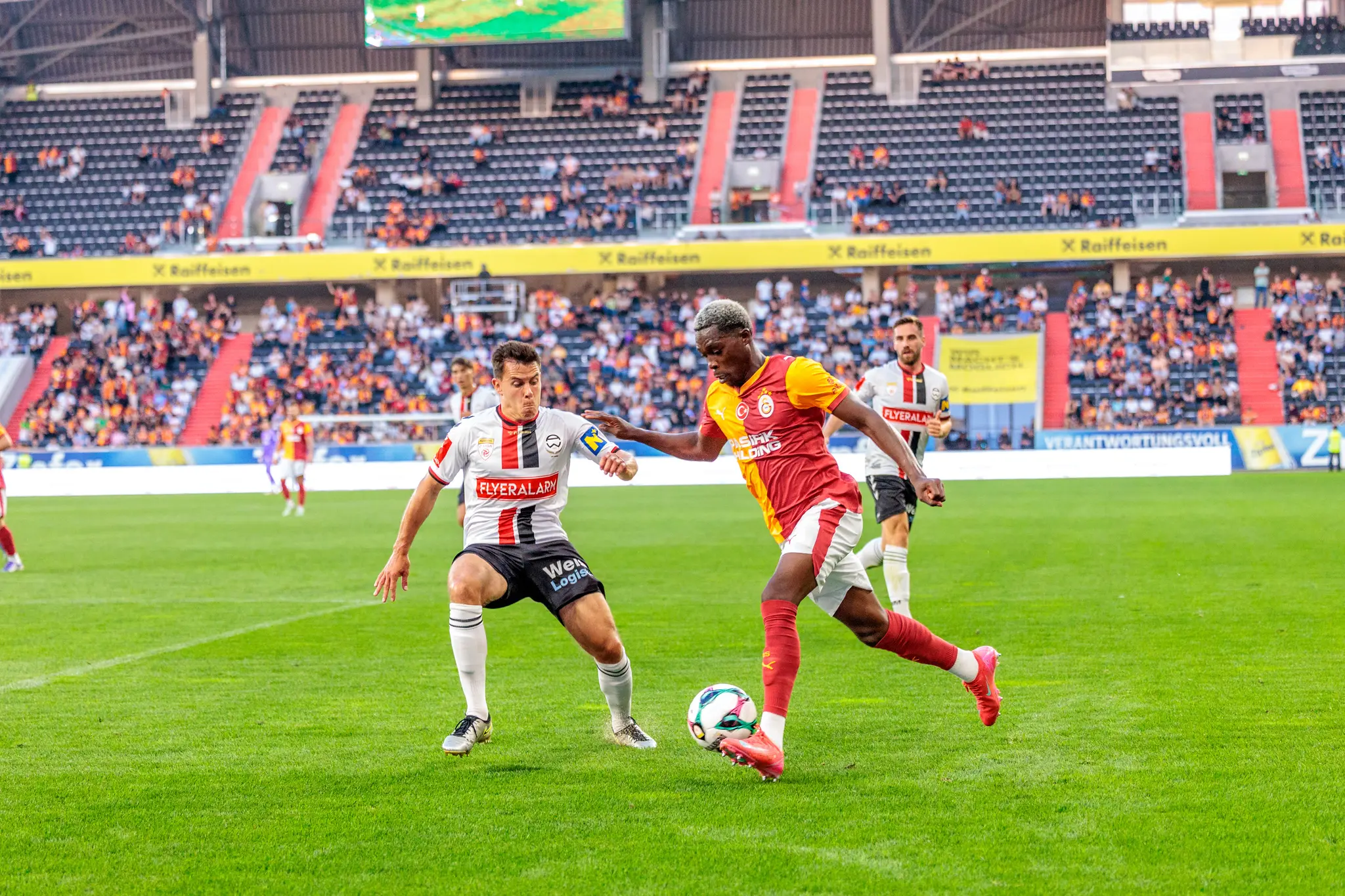 Zwei Fußballspieler, ein Admira Wacker und ein Galatasaray Spieler, duellieren sich im Spiel um den Ball in der Raiffeisen Arena vor Zuschauern 
